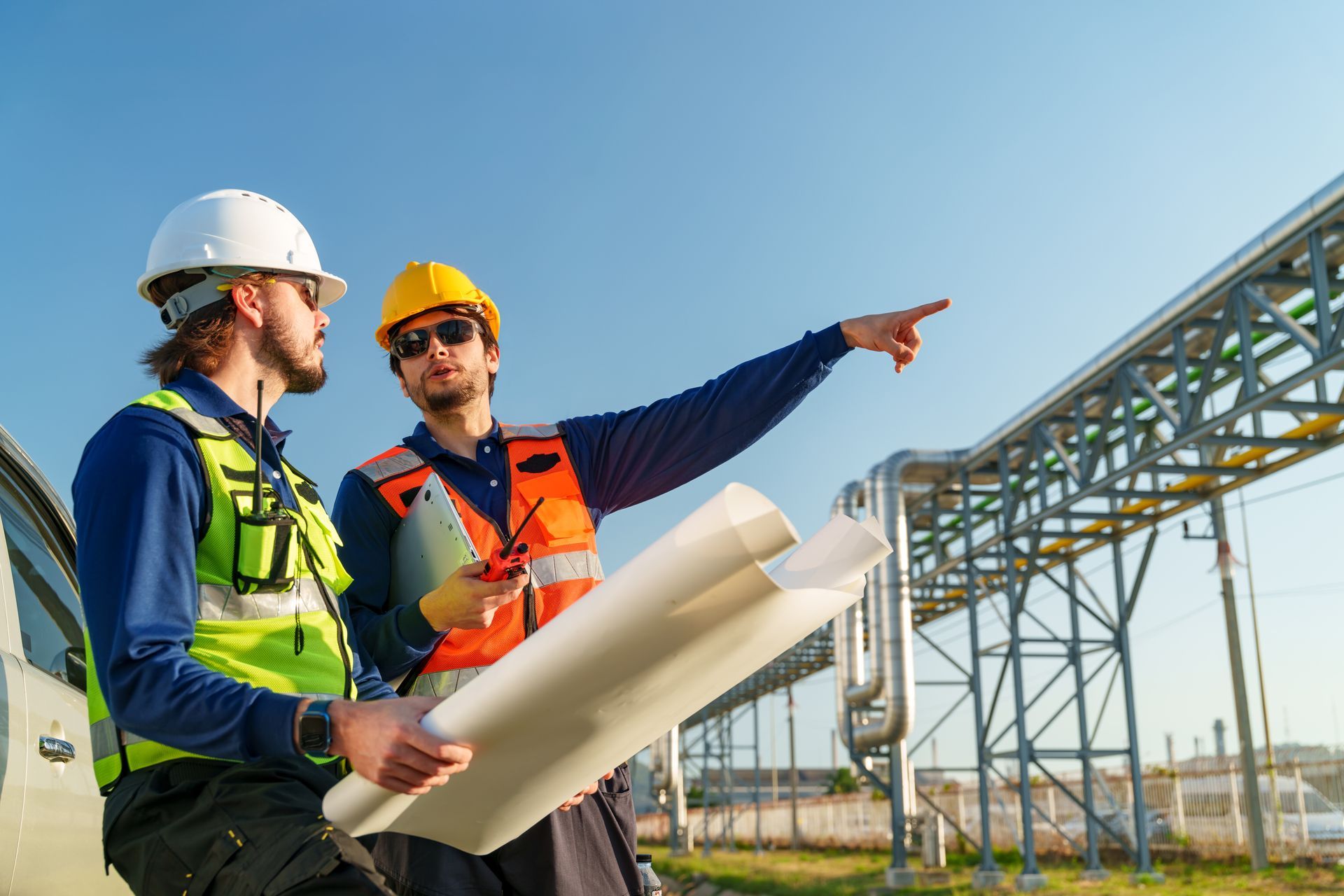 Two engineers in high-visibility vests and hard hats stand outdoors near industrial pipes, reviewing plans and pointing.
