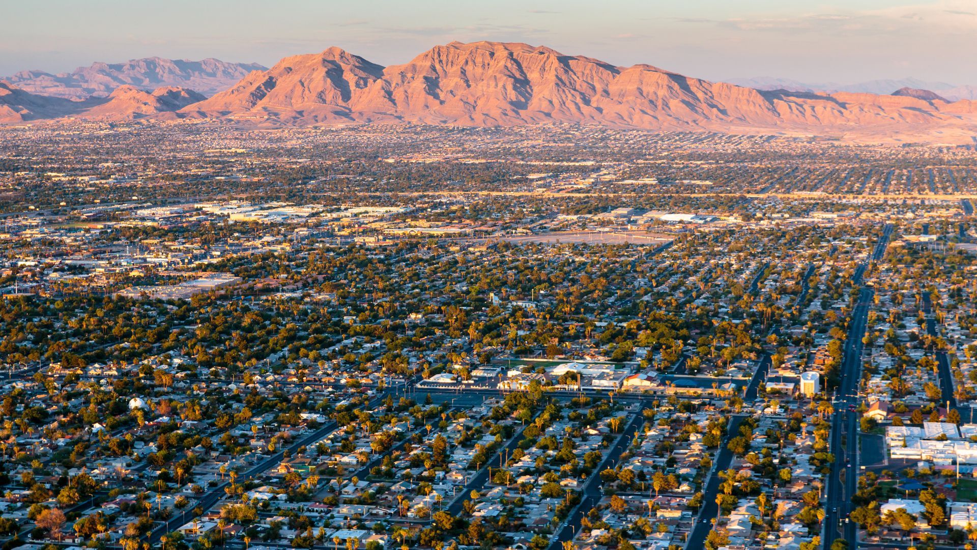 An aerial view of a sprawling desert city grid at golden hour, framed by pink-hued mountains under a clear sky.
