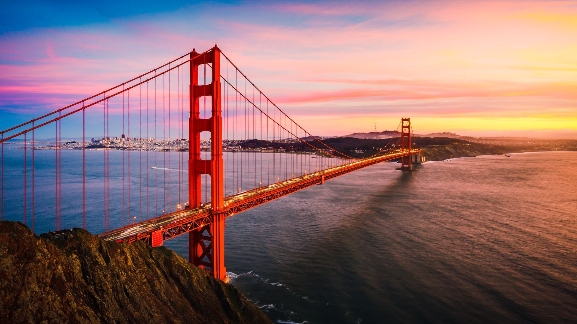 Golden Gate Bridge spanning the water at sunset with a pink and orange sky and city skyline in the distance.
