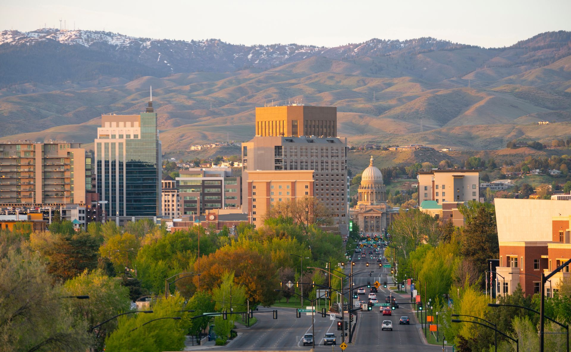 A city skyline at sunset with tall buildings, a prominent capitol dome, and foothills in the background.