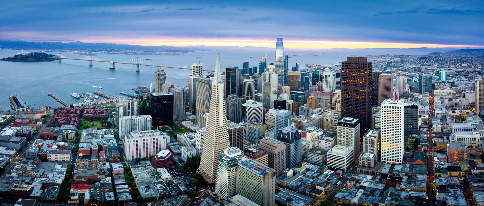 High-angle panoramic view of the San Francisco skyline at dusk, featuring the Transamerica Pyramid and the Bay Bridge.