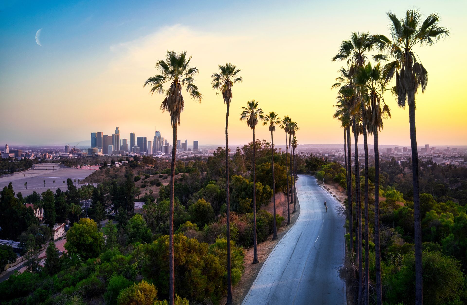 A road lined with tall palm trees at sunset, leading toward the Los Angeles city skyline in the distance.