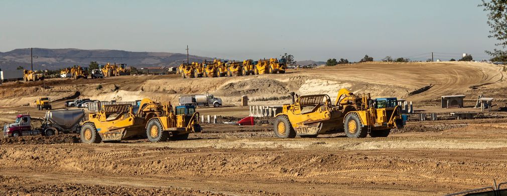 A wide-angle view of a construction site featuring multiple yellow earth-moving machines on a large, dry dirt terrain. A wide-angle view of a construction site featuring multiple yellow earth-moving machines on a large, dry dirt terrain.