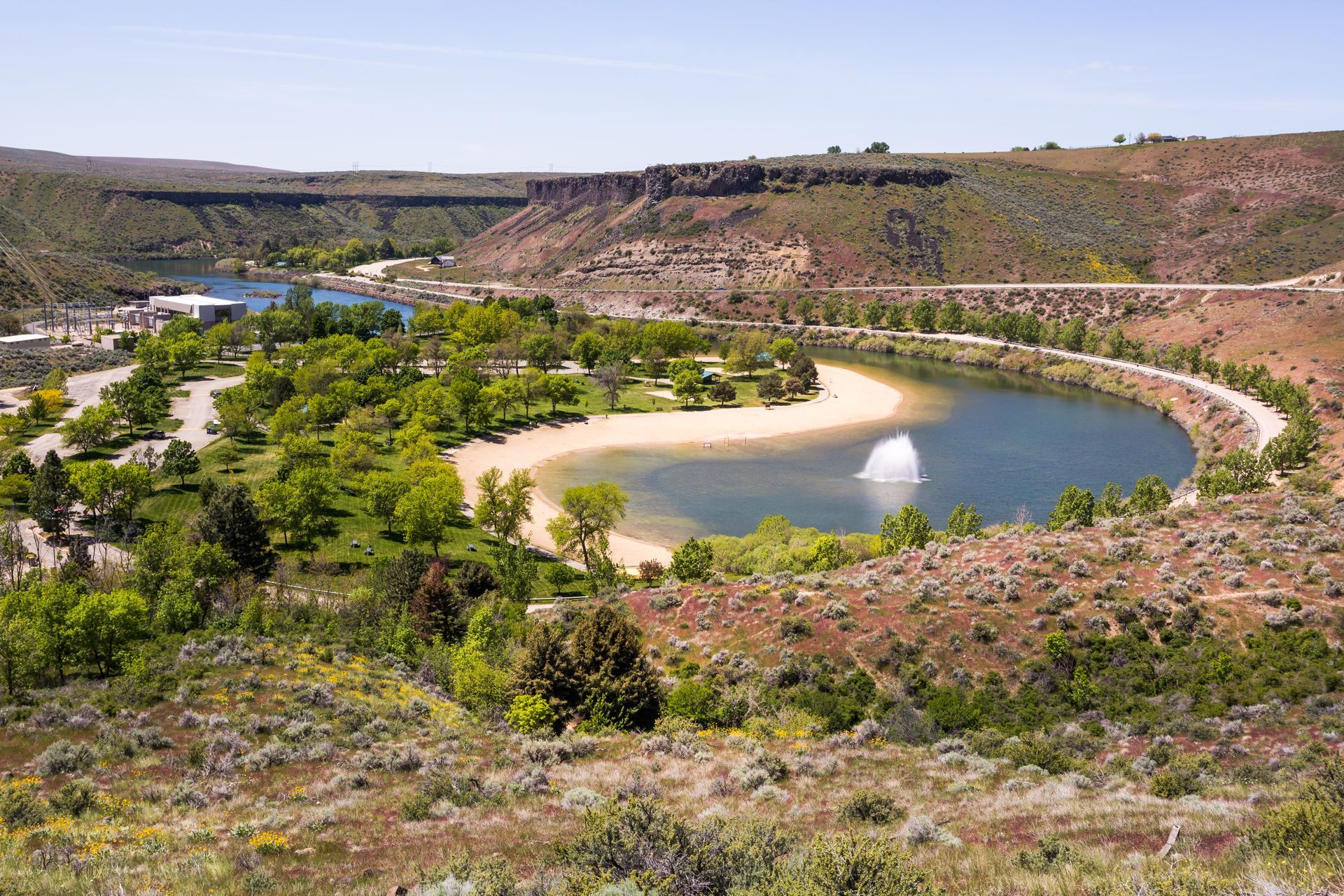 A fountain sprays water into a crescent-shaped lake surrounded by trees, a beach, and rocky canyon walls.