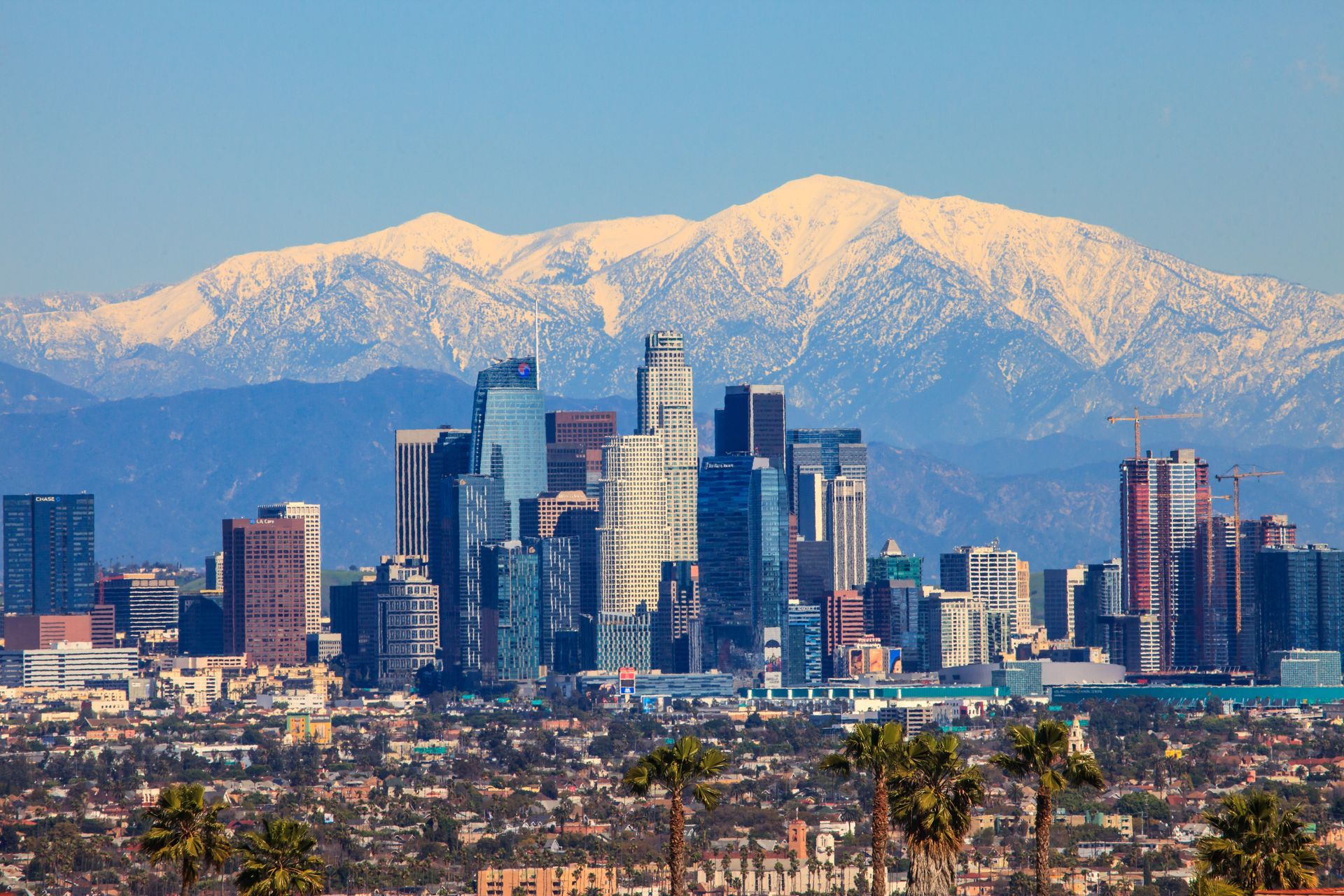 The Los Angeles skyline under a clear blue sky, set against a backdrop of snow-capped mountains.
