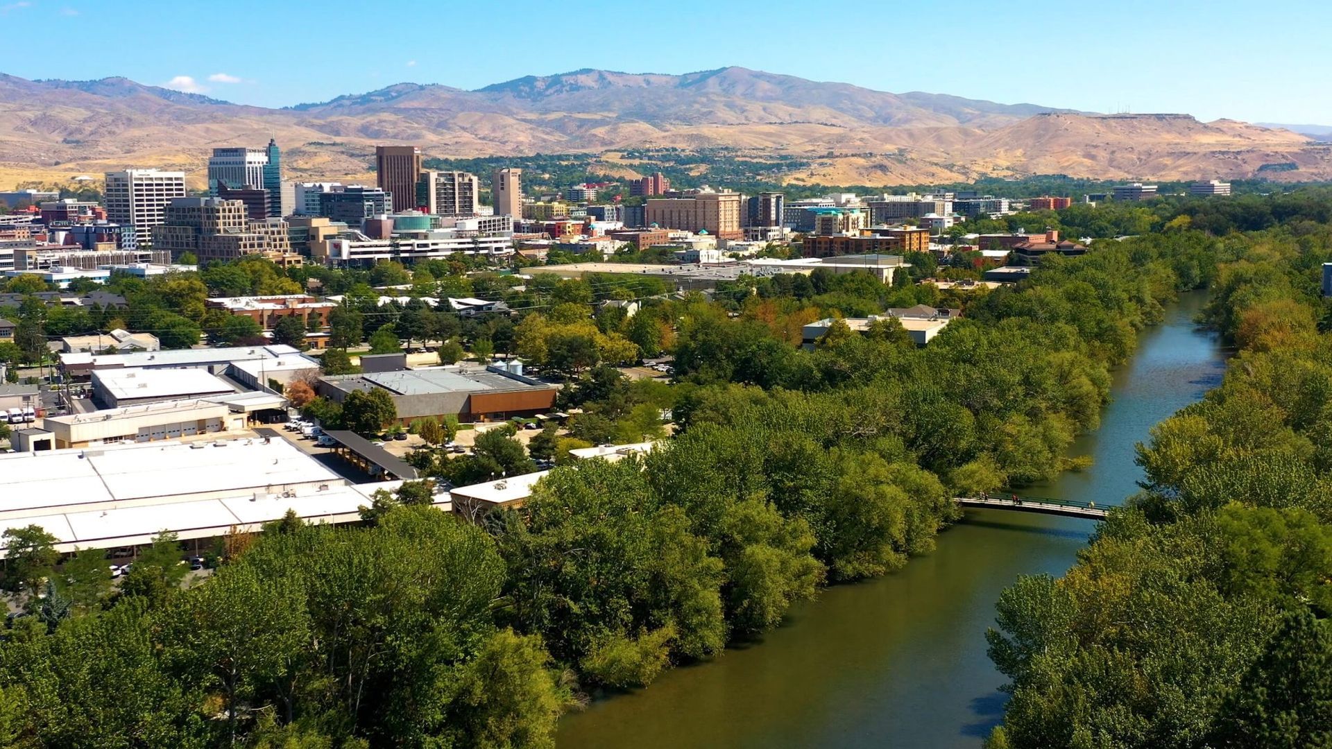 A Boise, Idaho city skyline with the Boise River in the foreground, surrounded by trees under a clear blue sky.