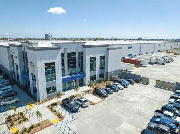 Modern, multi-story industrial warehouse facility with blue accents, a parking lot, and delivery trucks on a sunny day.