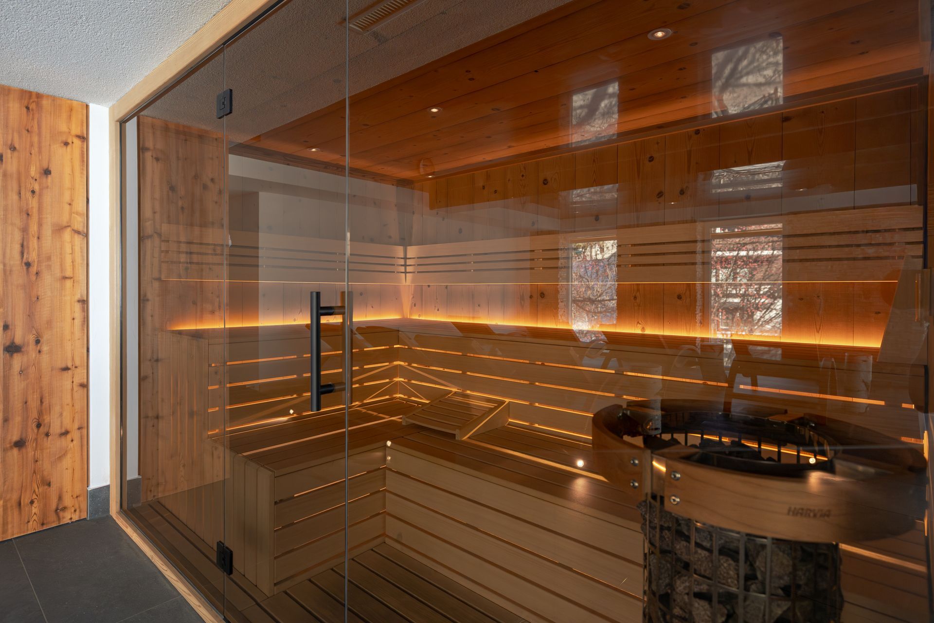 Modern sauna with wood and glass walls, warm amber lighting, and a round heater bench inside.
