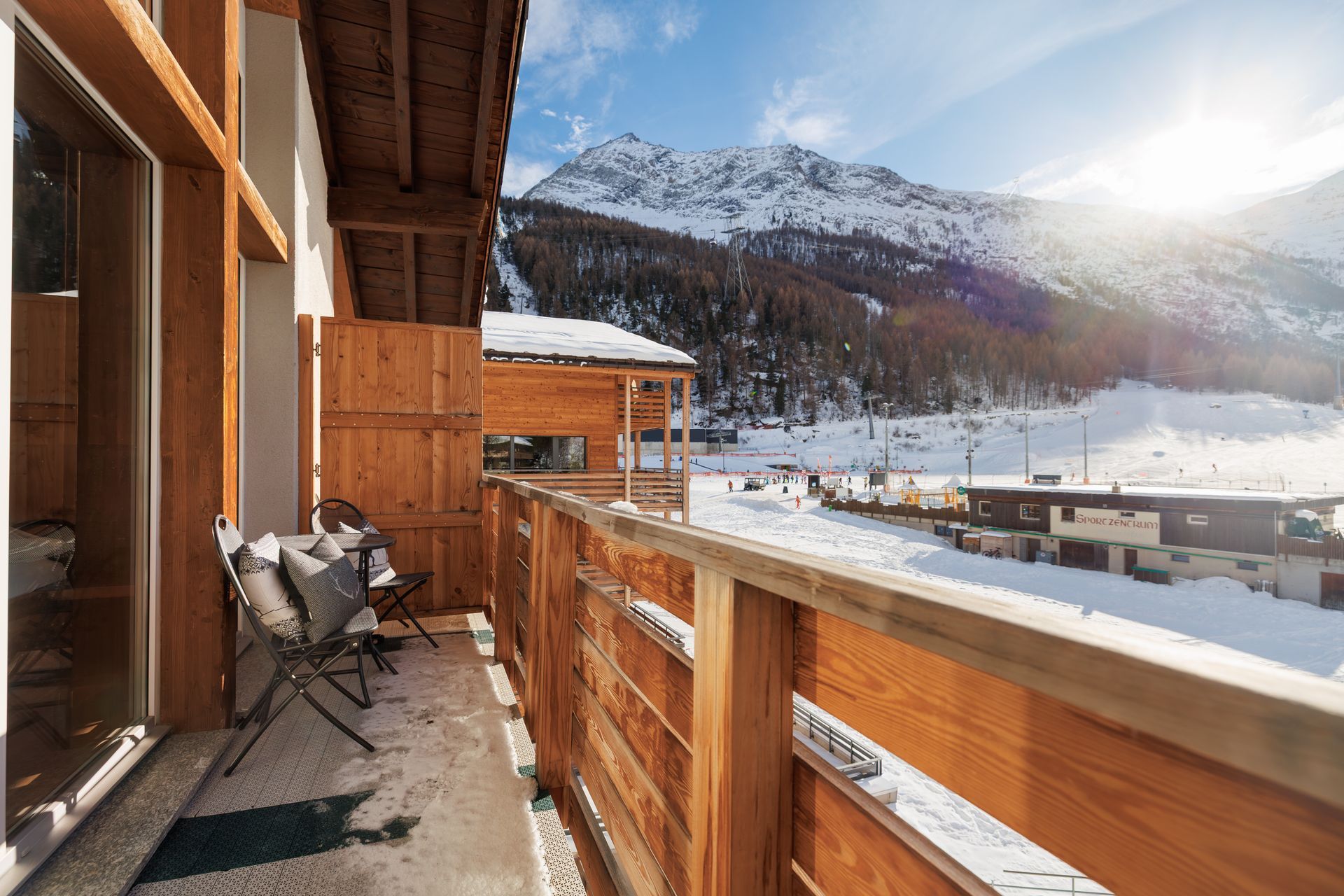 Snowy mountain view from a wooden balcony with chairs and bright winter sunlight.