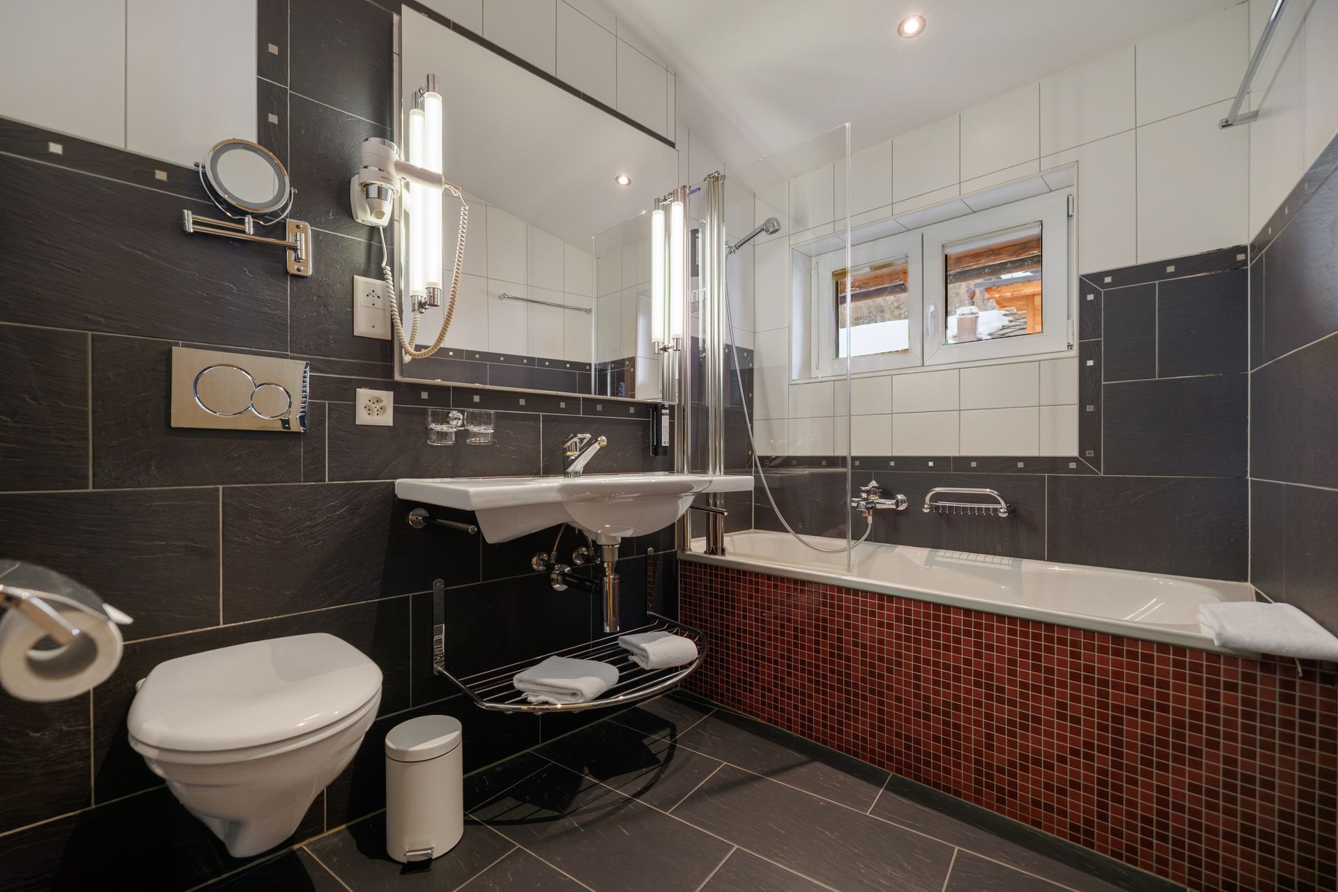 Modern bathroom with black tile, white sink and toilet, large mirror, and a bathtub under a window