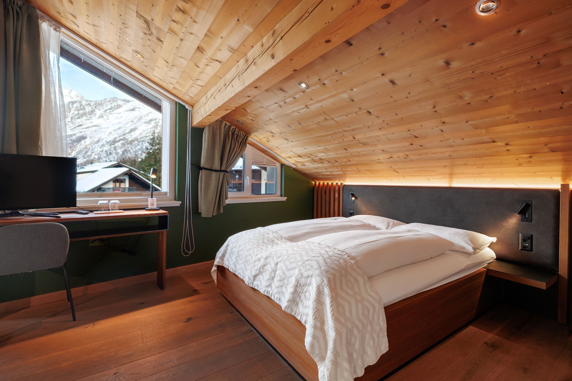 Cozy attic bedroom with sloped wooden ceiling, bed, desk, and large windows overlooking trees.