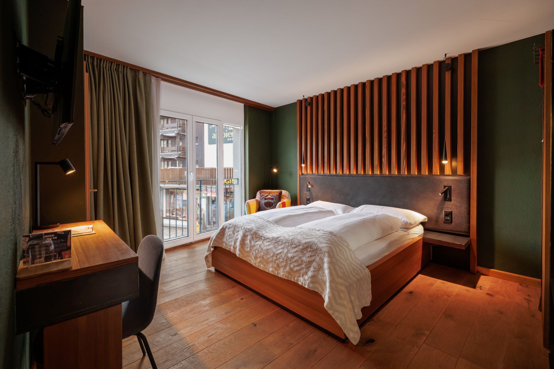 Cozy modern bedroom with a large bed, warm wood accents, desk, and balcony doors with city view