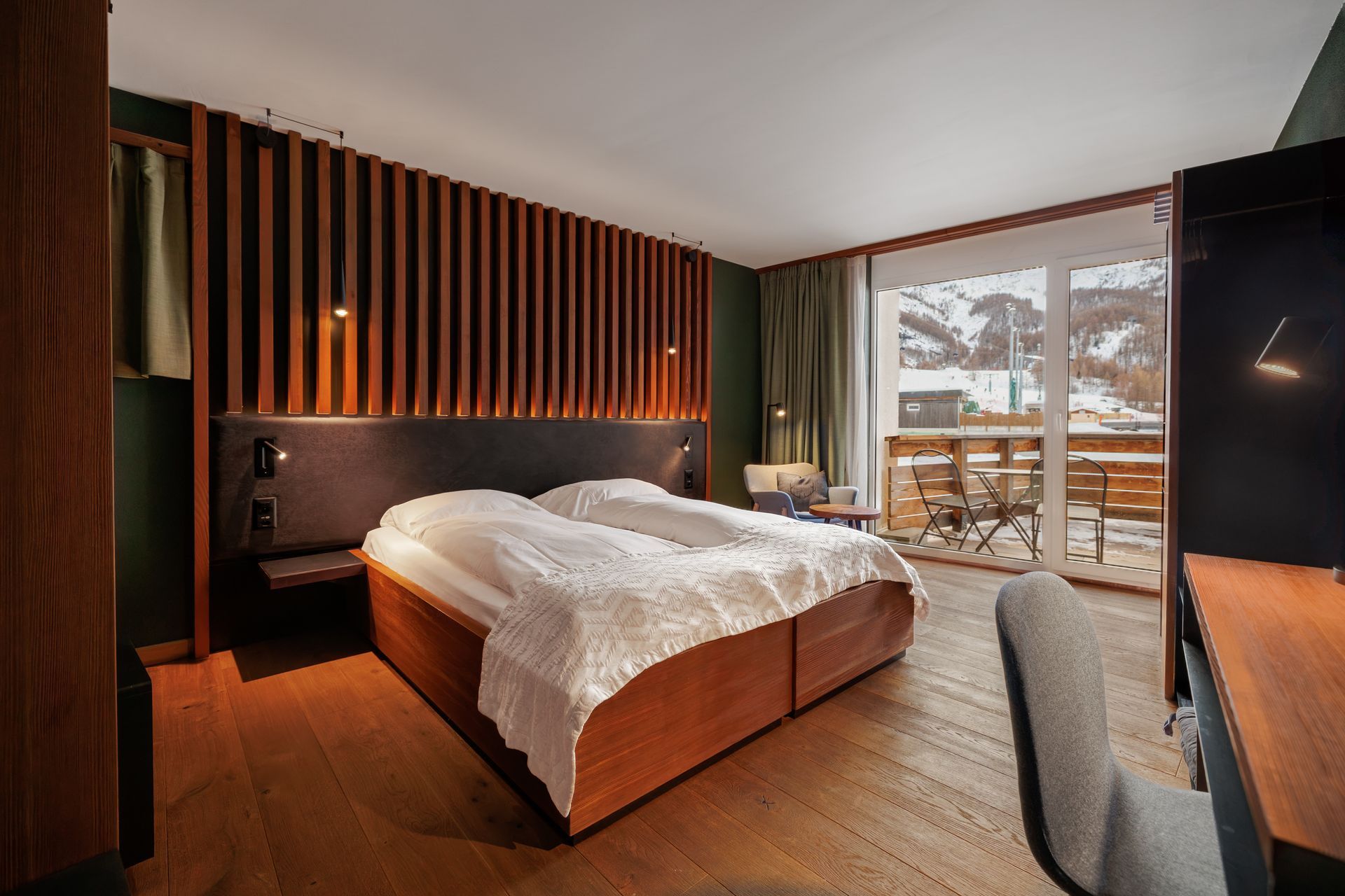 Modern bedroom with wooden bed, striped accent wall, warm lighting, and large balcony windows.