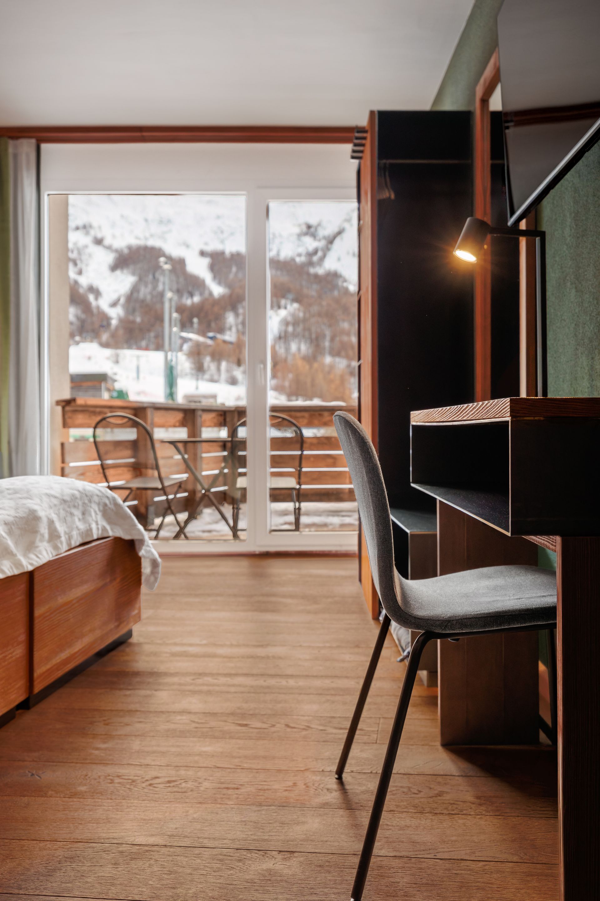 Cozy bedroom with wooden floor, bed, desk chair, and balcony doors overlooking snowy mountains