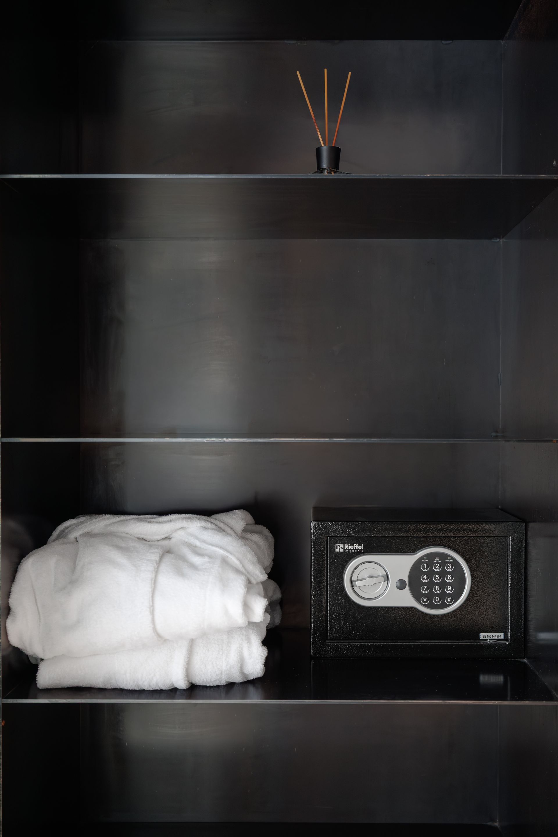 Dark shelf with folded white towels, a small diffuser on top, and a black safe on the lower shelf