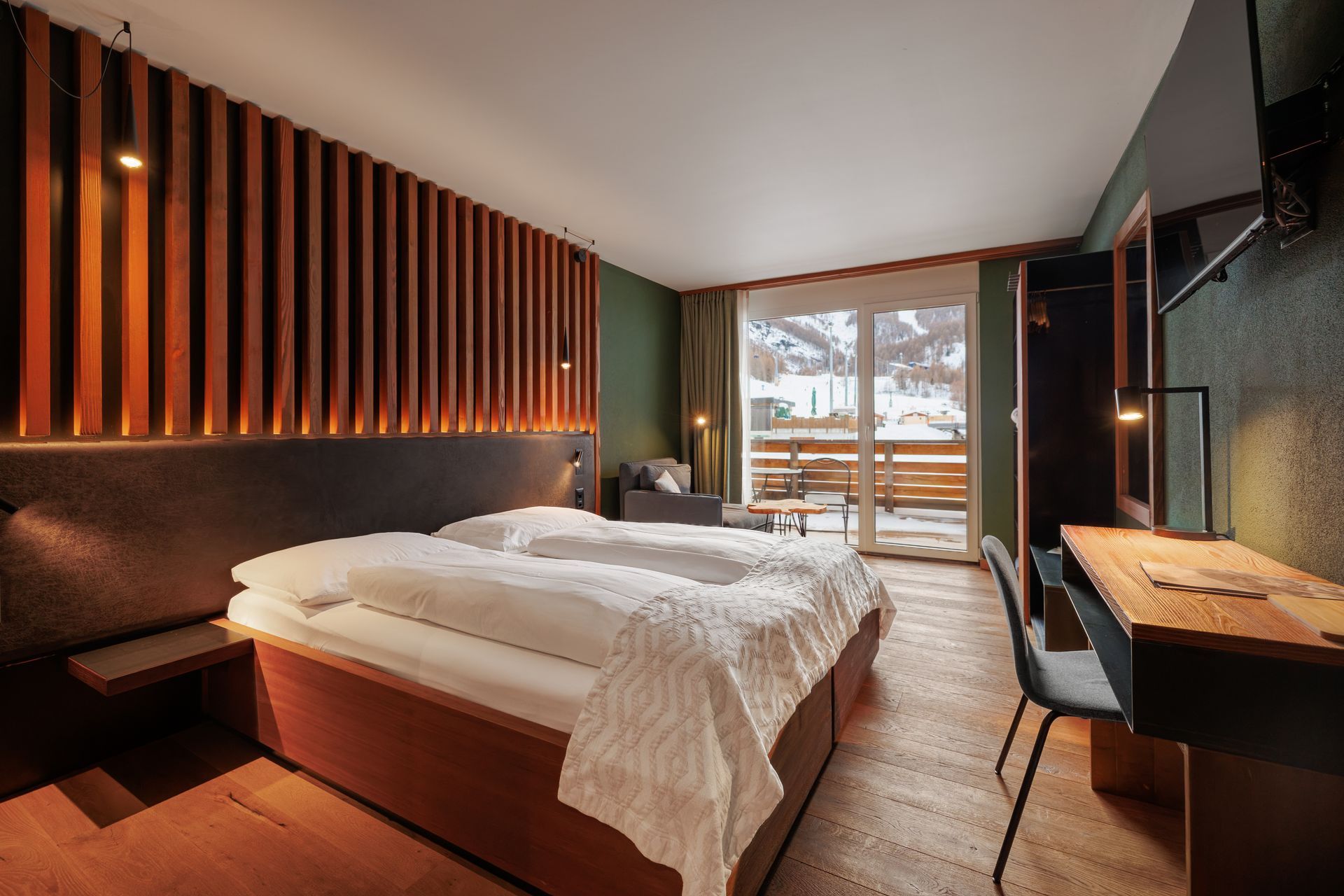 Modern hotel room with bed, wood accents, desk, and large window overlooking snow