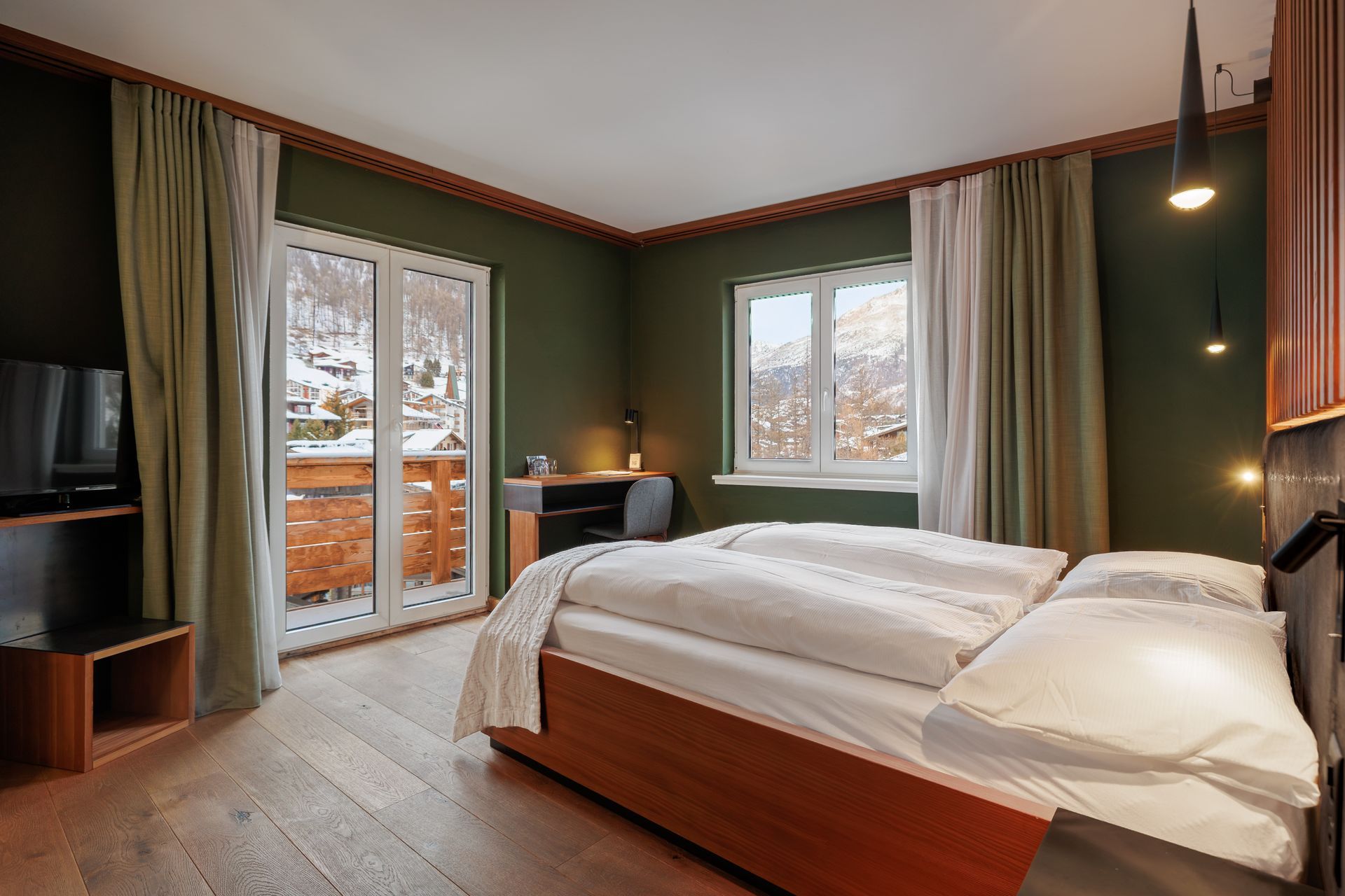 Modern bedroom with a wooden bed, green walls, windows, and a balcony door overlooking snowy trees