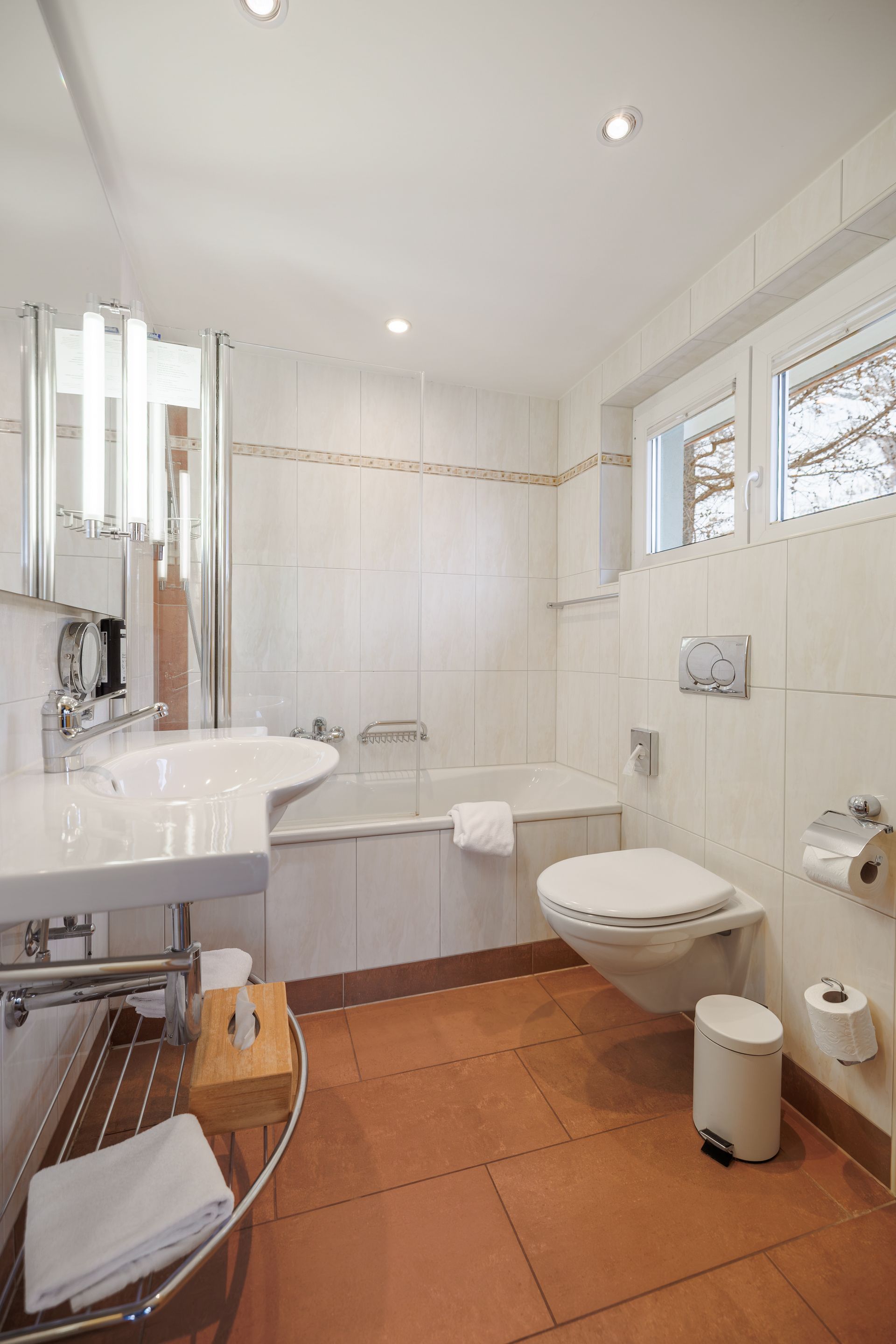 Bright white bathroom with bathtub, sink, toilet, orange tile floor, and small window.