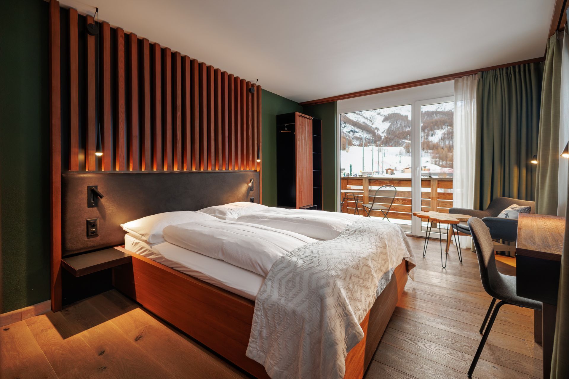 Modern hotel bedroom with a large bed, wood slat accent wall, and balcony doors overlooking a winter view