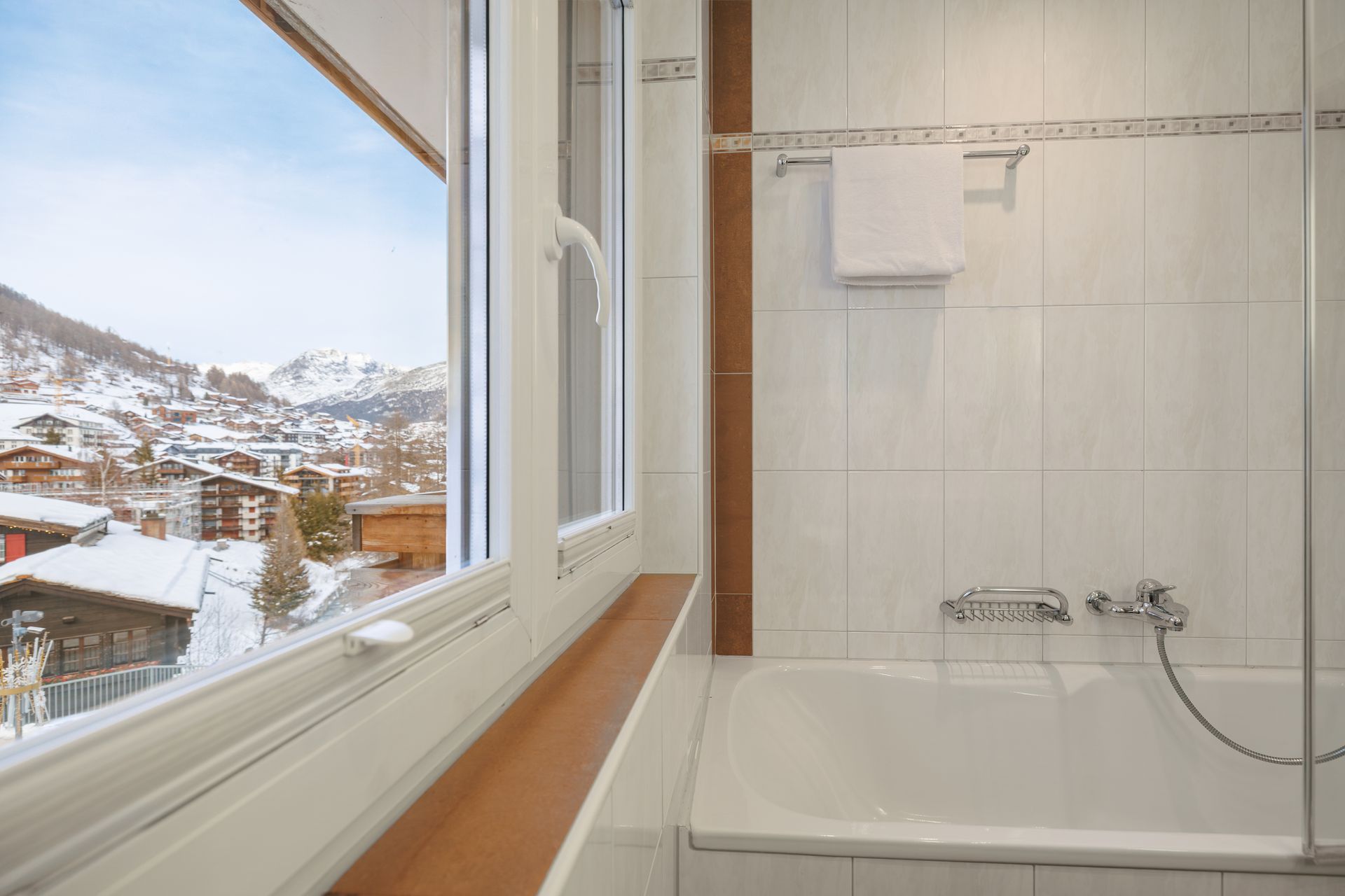 Bright bathroom with a bathtub beside a window overlooking snowy houses and mountains.