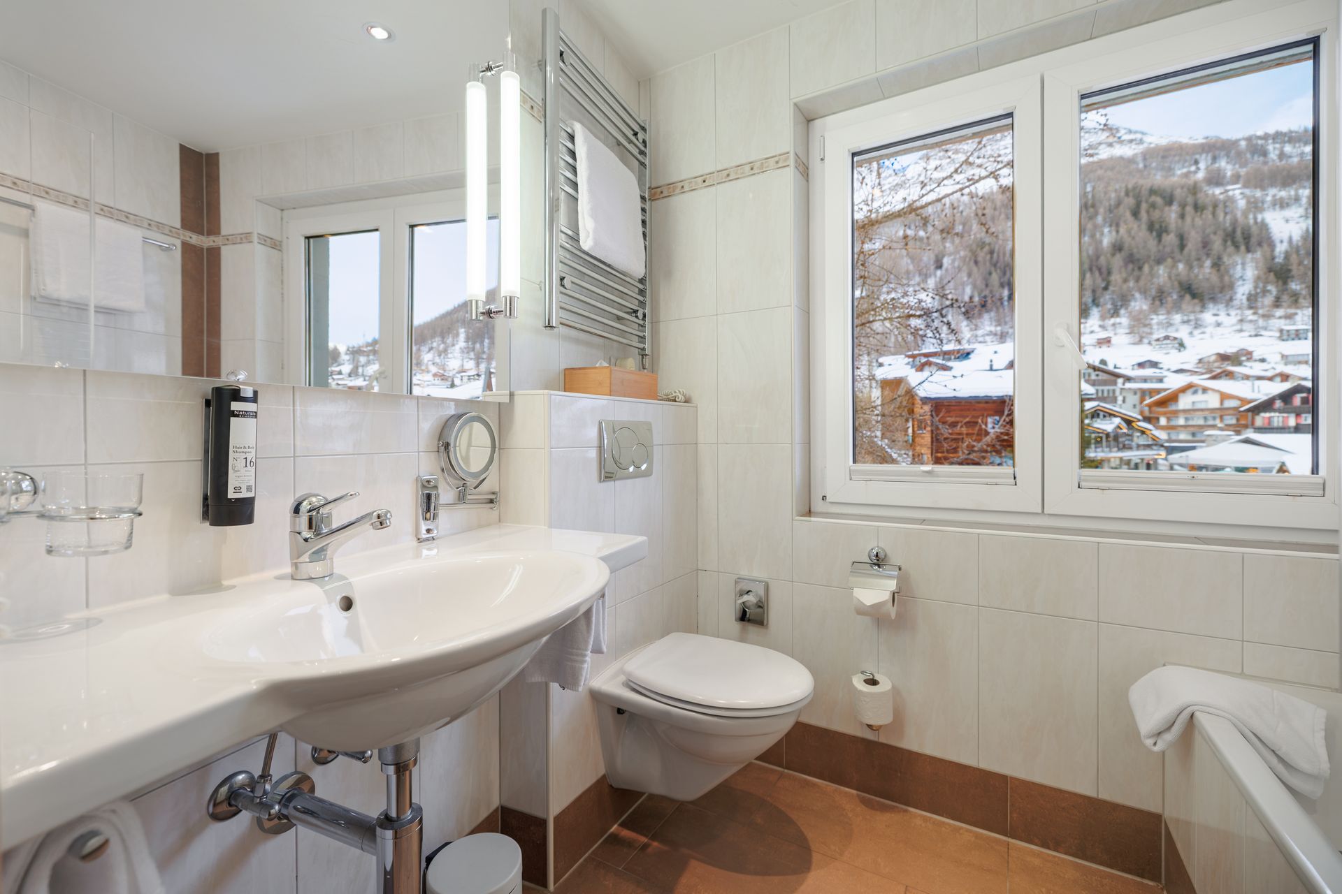 Bright bathroom with sink, toilet, towel rack, and snowy mountain view through the window.