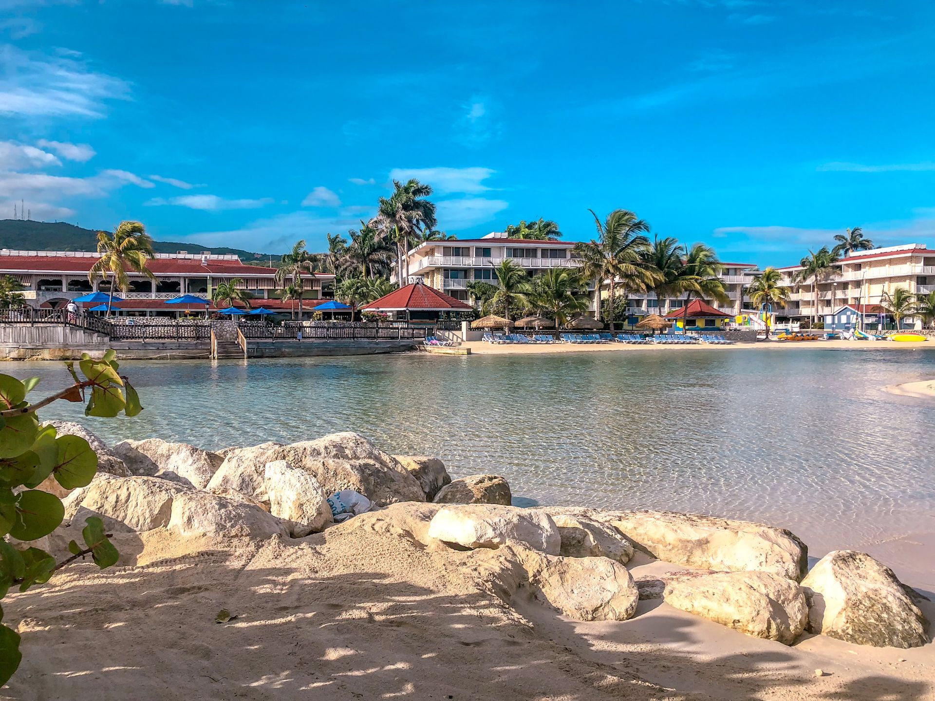 Rocky shoreline overlooking a calm bay with beachside buildings, palm trees, and a bright blue sky