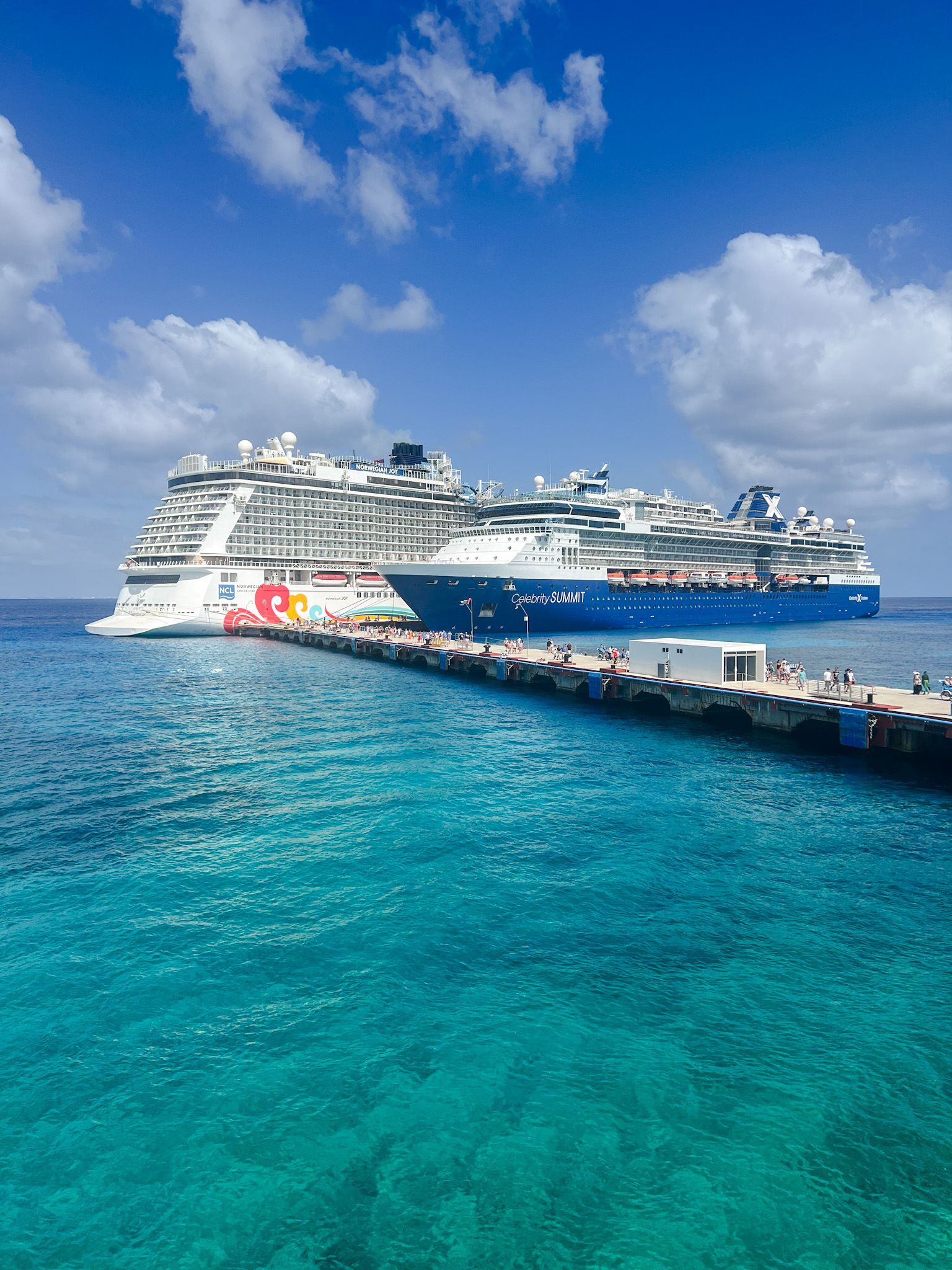 Two large cruise ships docked beside a turquoise sea under a blue sky with clouds.