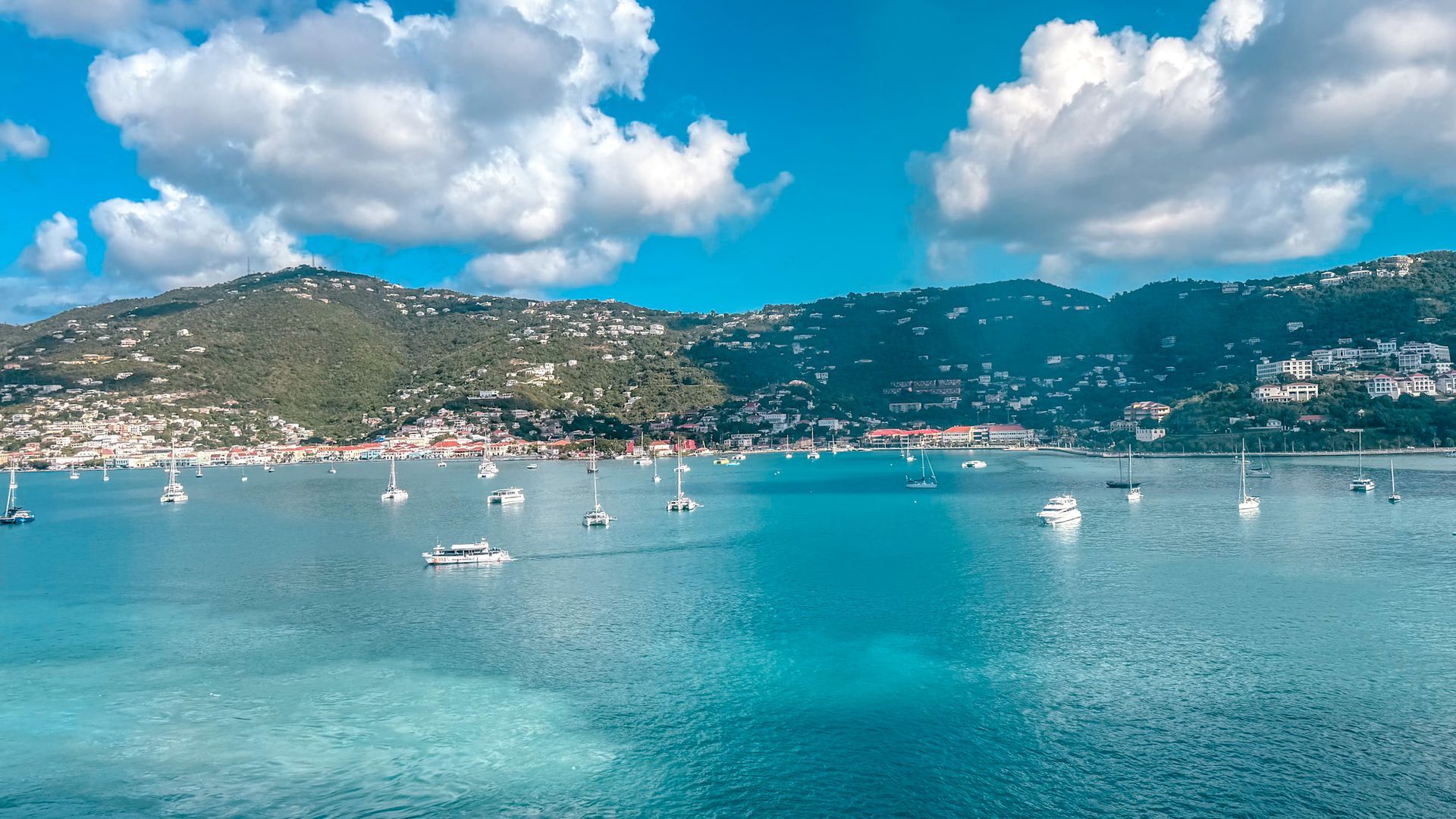 Calm turquoise bay with sailboats, a hillside town, and bright clouds in a blue sky