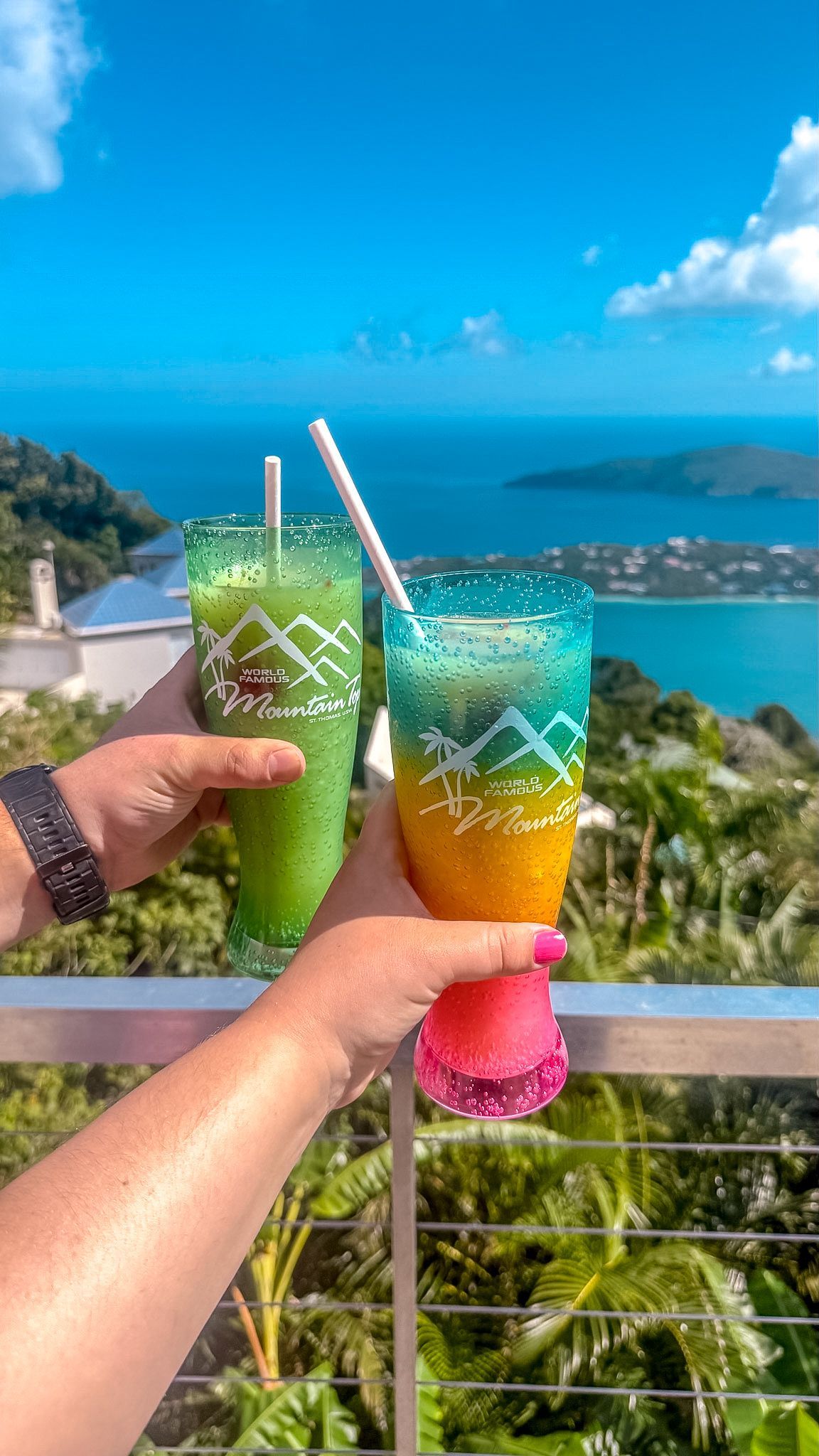 Two colorful tropical drinks raised against a sunny ocean and island view from a balcony