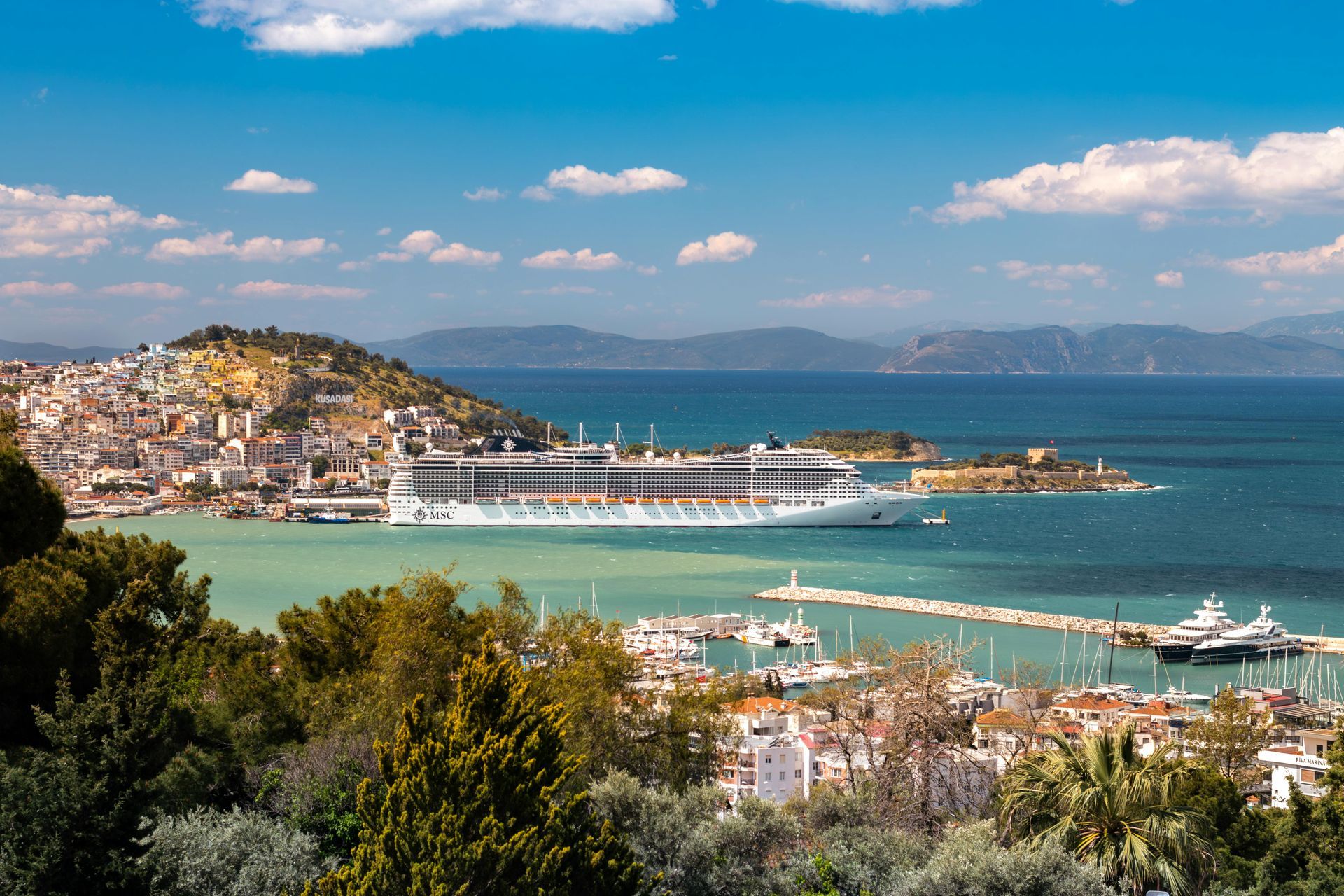 Coastal harbor town with white buildings, turquoise water, yachts, and hills under a sunny sky