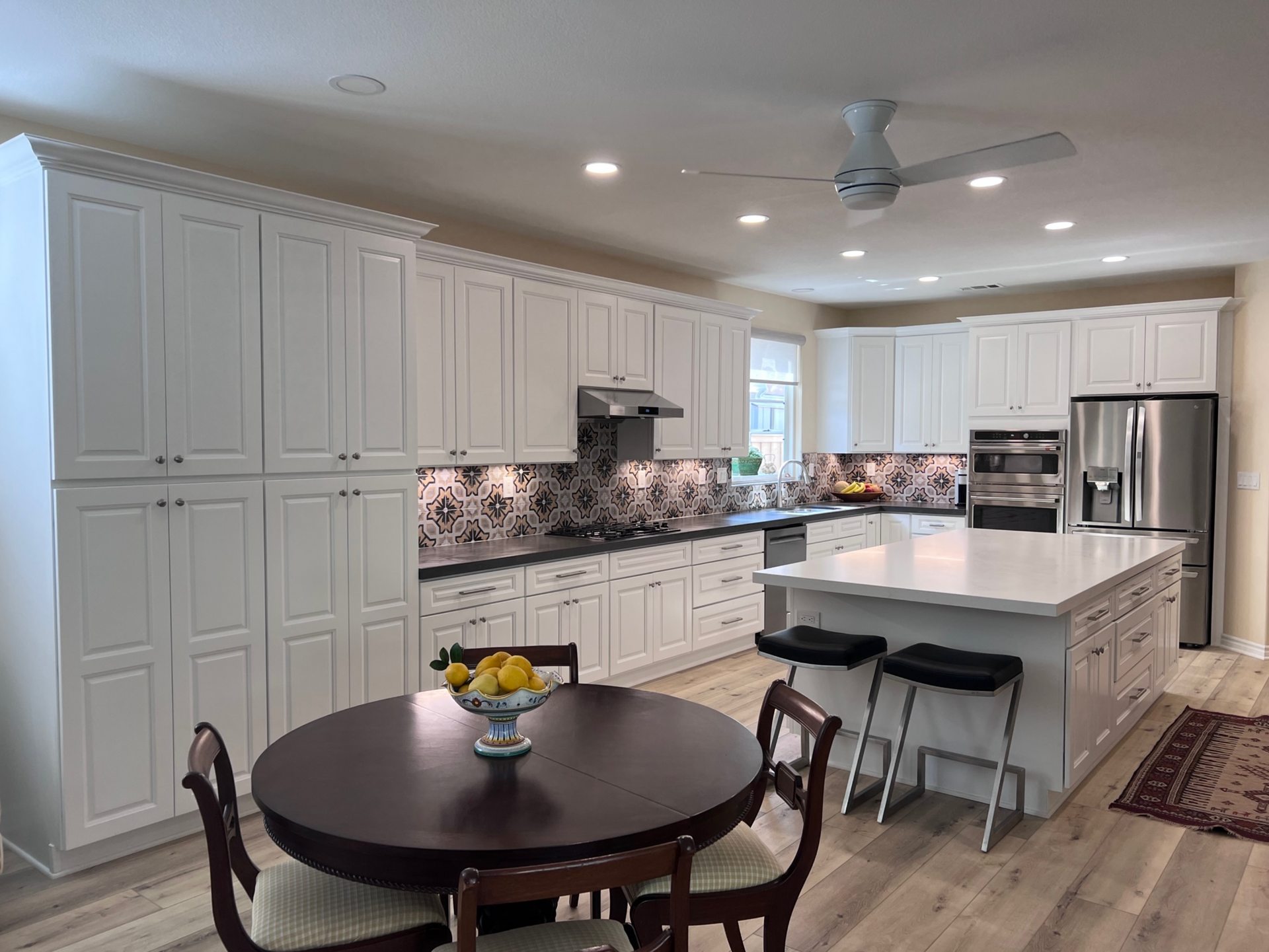 A kitchen with white cabinets , stainless steel appliances , a large island , a table and chairs.
