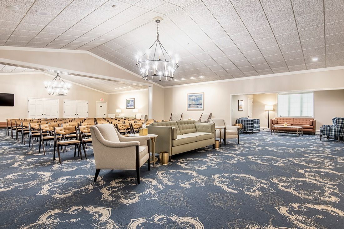 Spacious hall with blue patterned carpet, a white sofa and armchairs, and rows of chairs. A chandelier hangs from the ceiling.