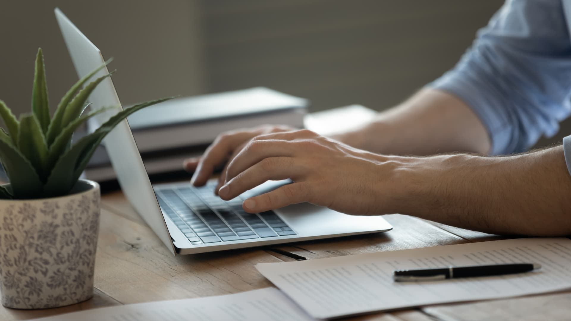 Person typing on a laptop with hands on the keyboard. A pen and paper, a plant, and books are on the desk.