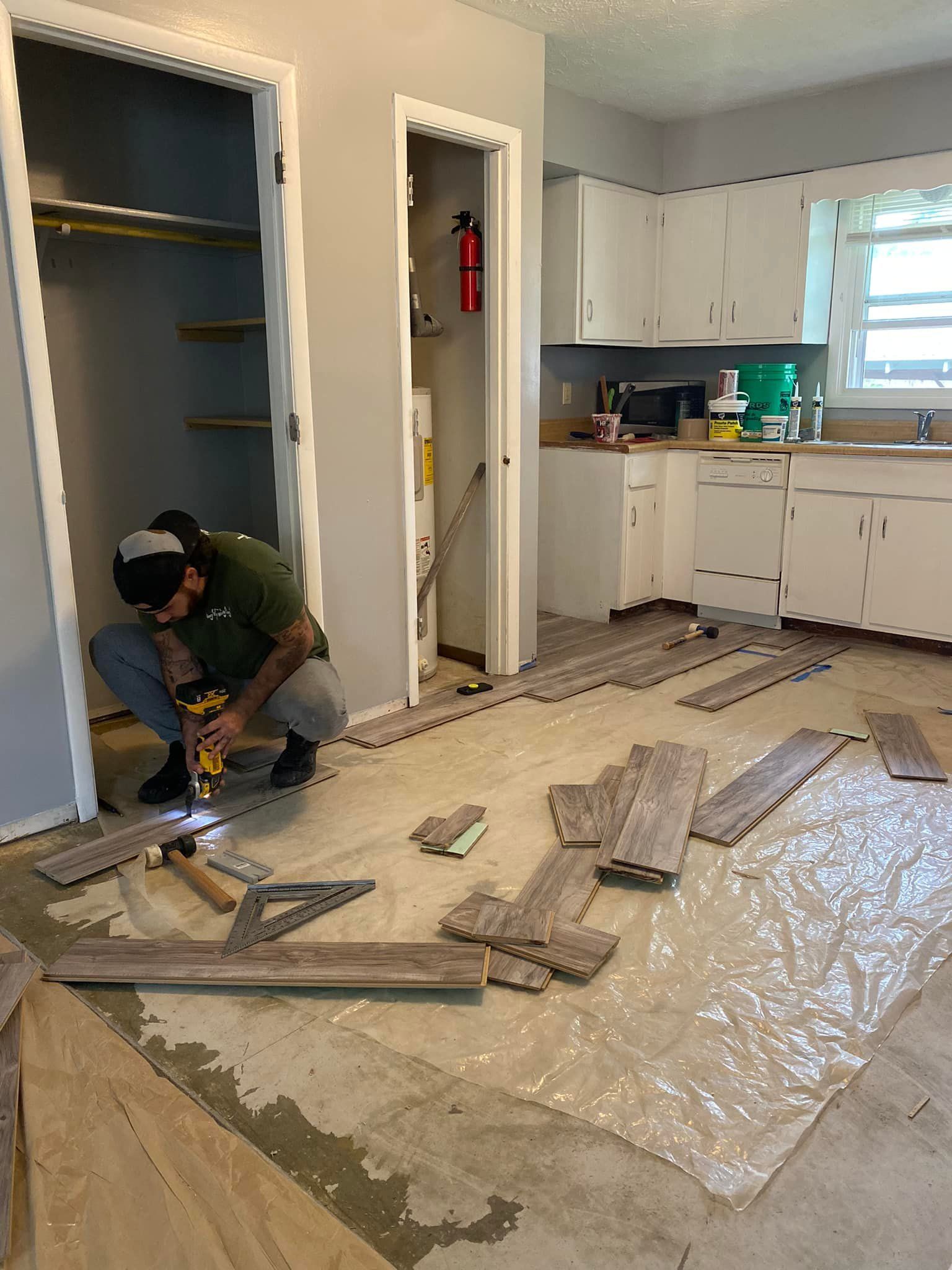 Man installing floorboards in a room with a closet and kitchen. Gray walls, light wood-look flooring.