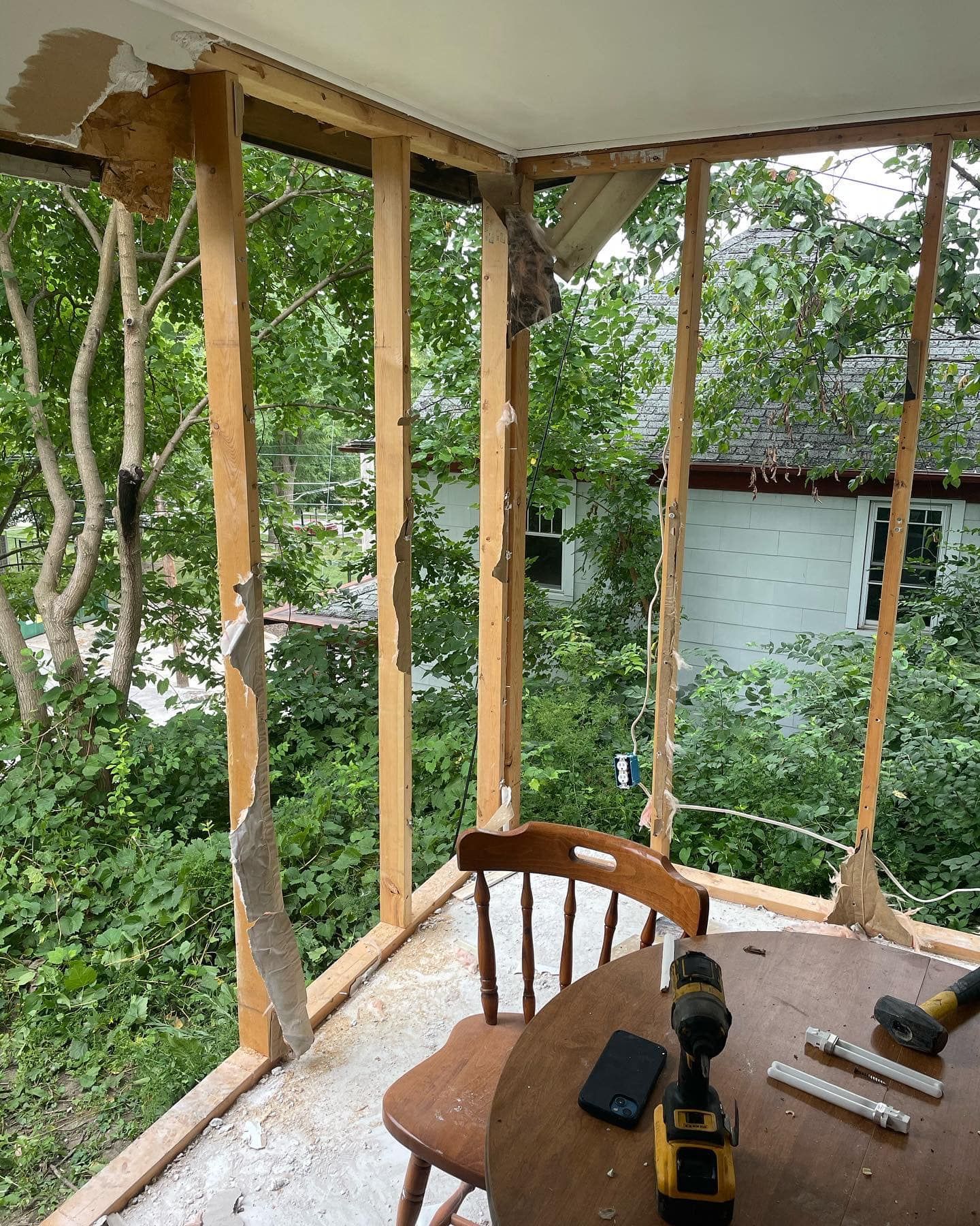 Wooden framed room open to the outside with a table, chair, and tools inside. Trees and a house are visible.
