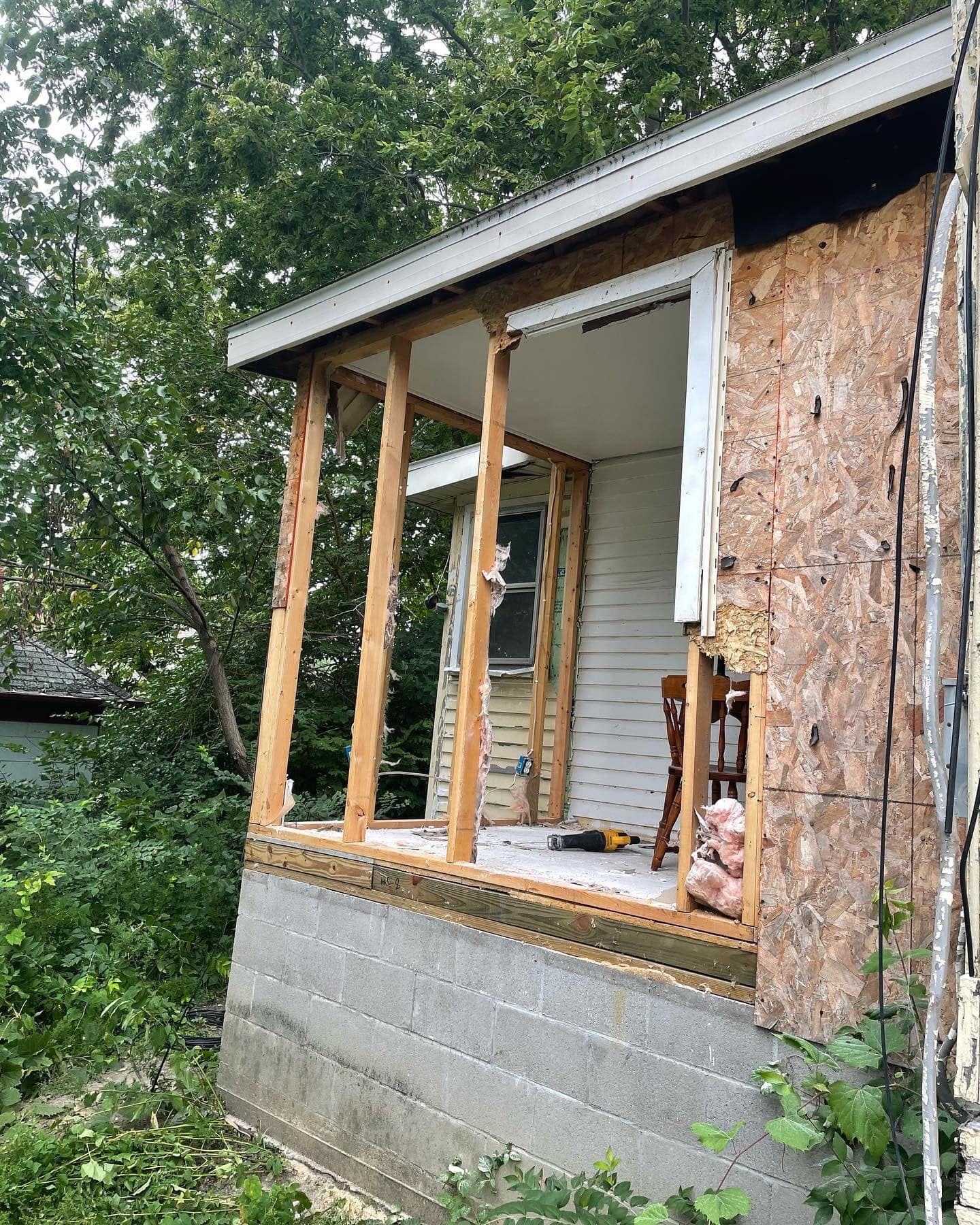 Porch demolition; plywood siding, exposed framing, gray foundation. Overgrown vegetation surrounds.