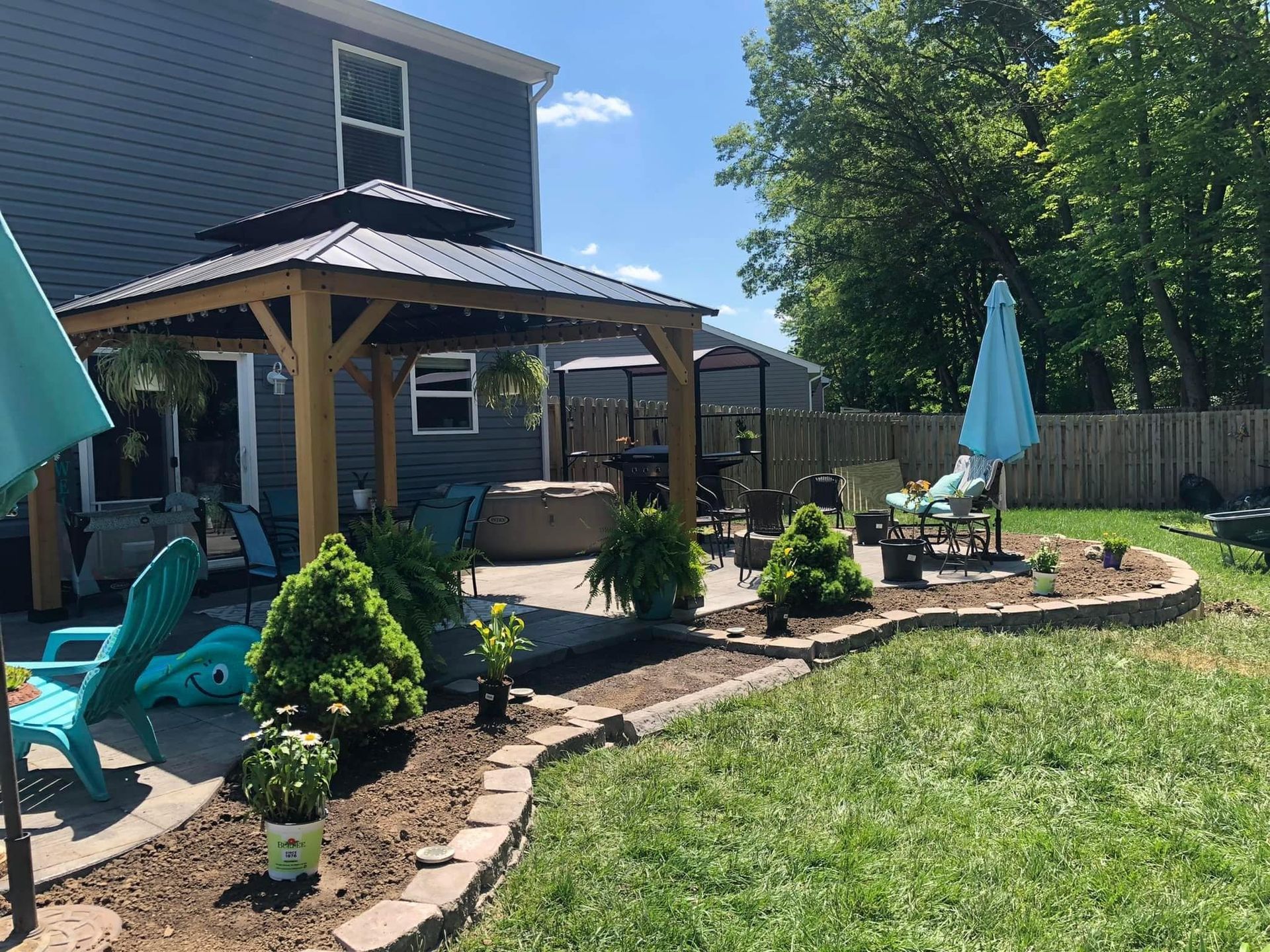 Backyard patio with gazebo, plants, seating, and blue umbrellas, on a sunny day.