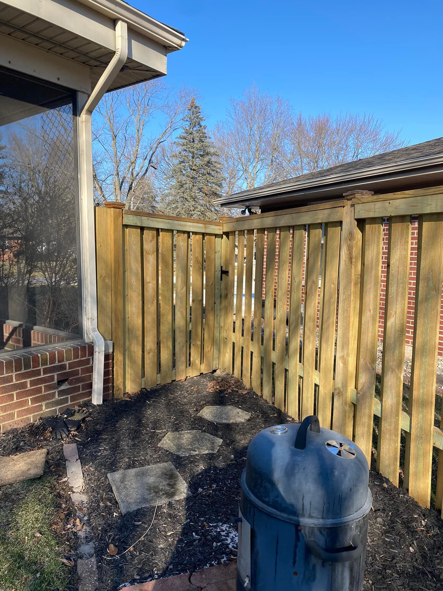 Wooden fence next to a brick wall, with a grill, stone steps, and trees in the background.