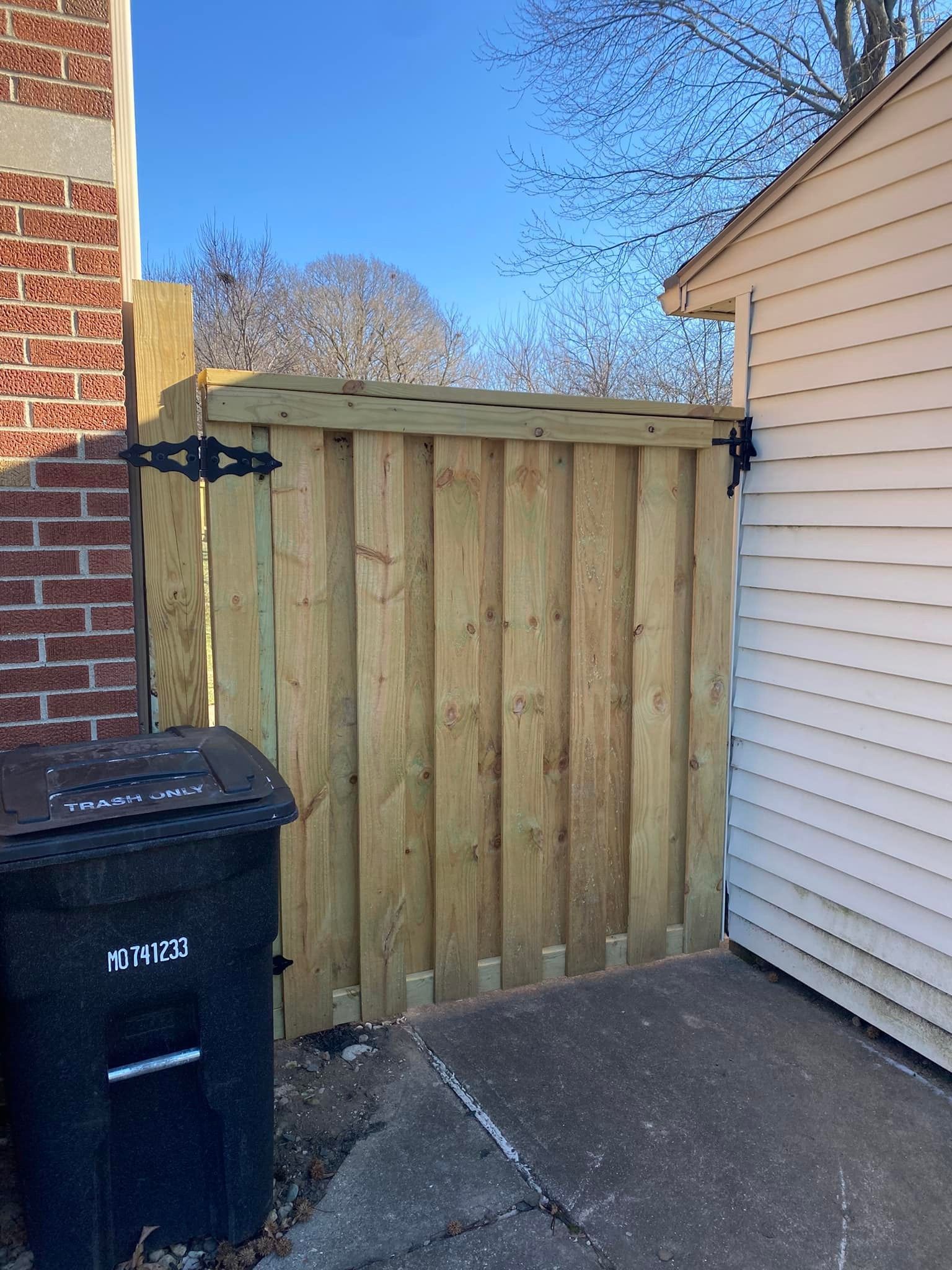 Wooden gate between brick wall and white siding, with black hinges and a trash can.
