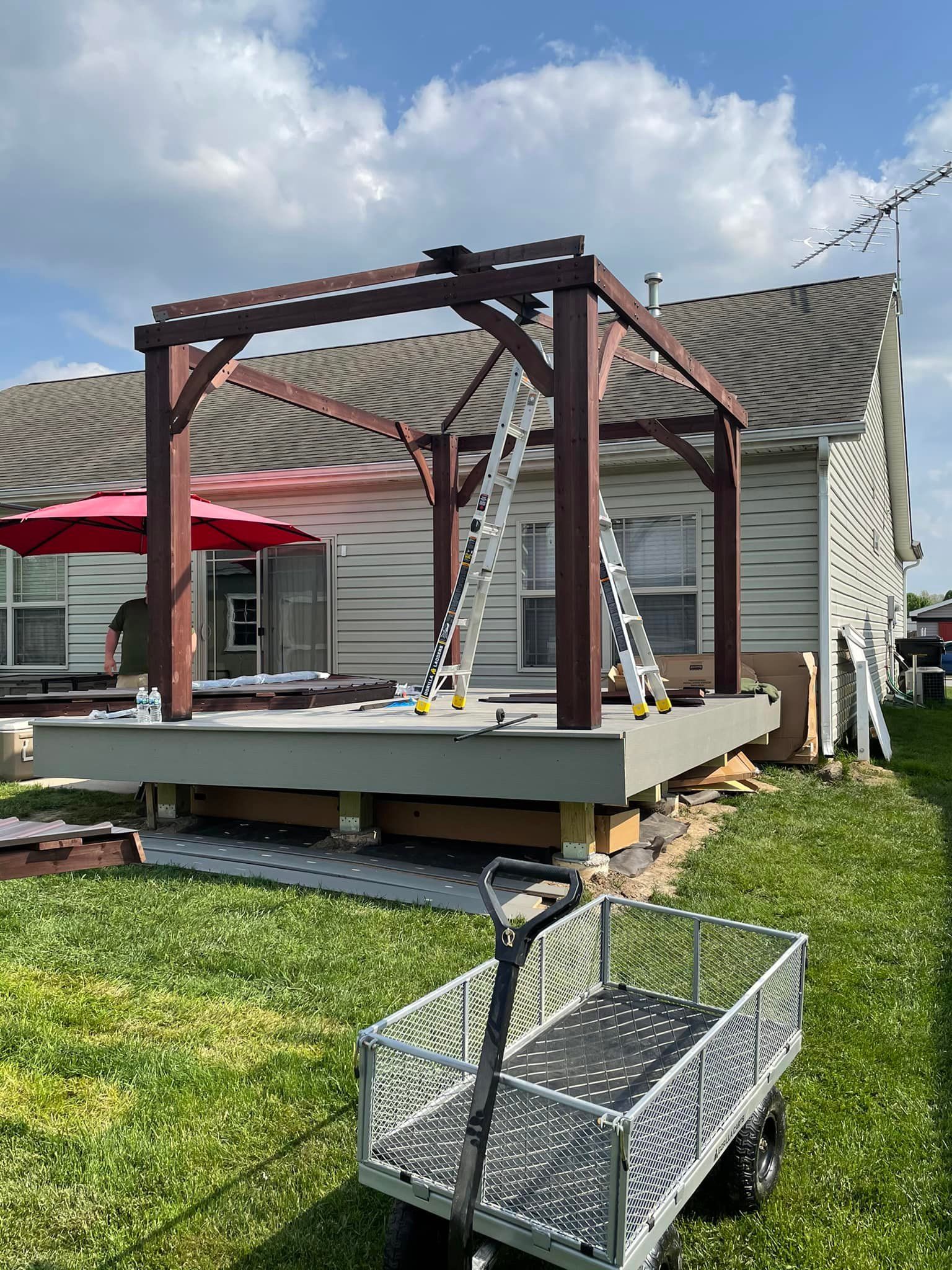 Pergola under construction on a deck with a ladder, wagon, and house in the background on a sunny day.