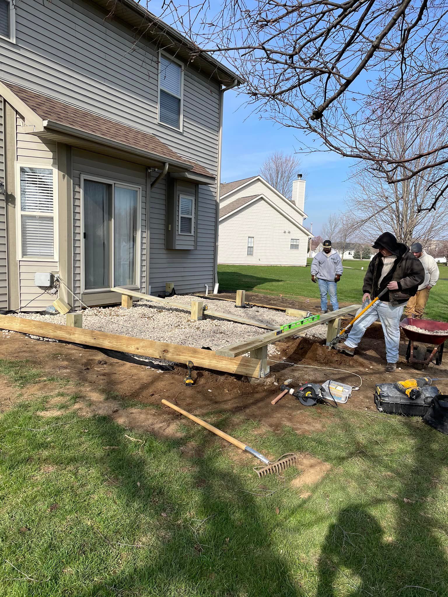 Construction crew building a deck next to a two-story house on a sunny day. One man shovels dirt.