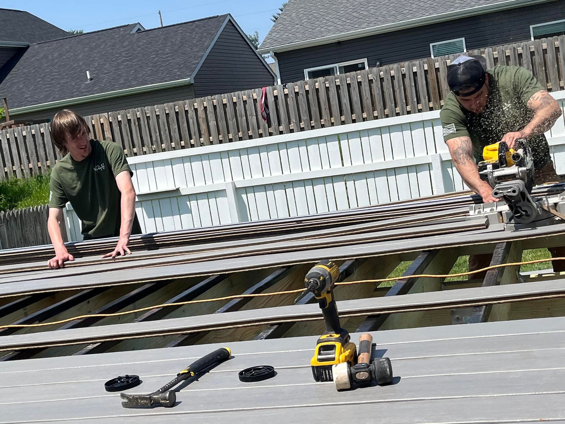 Two men building a deck on a sunny day. One uses a saw, the other works on boards.