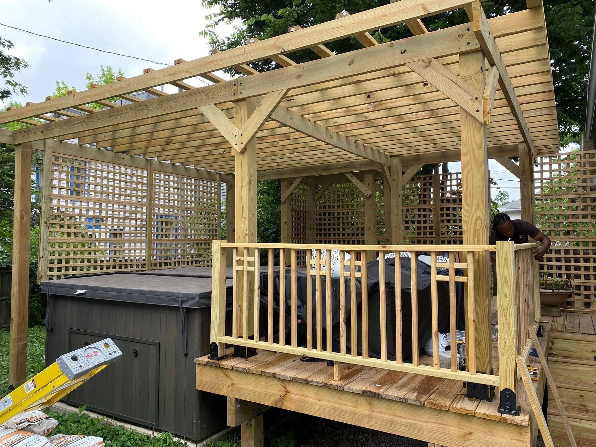Wooden pergola over a hot tub on a deck with lattice and railings, under a cloudy sky.