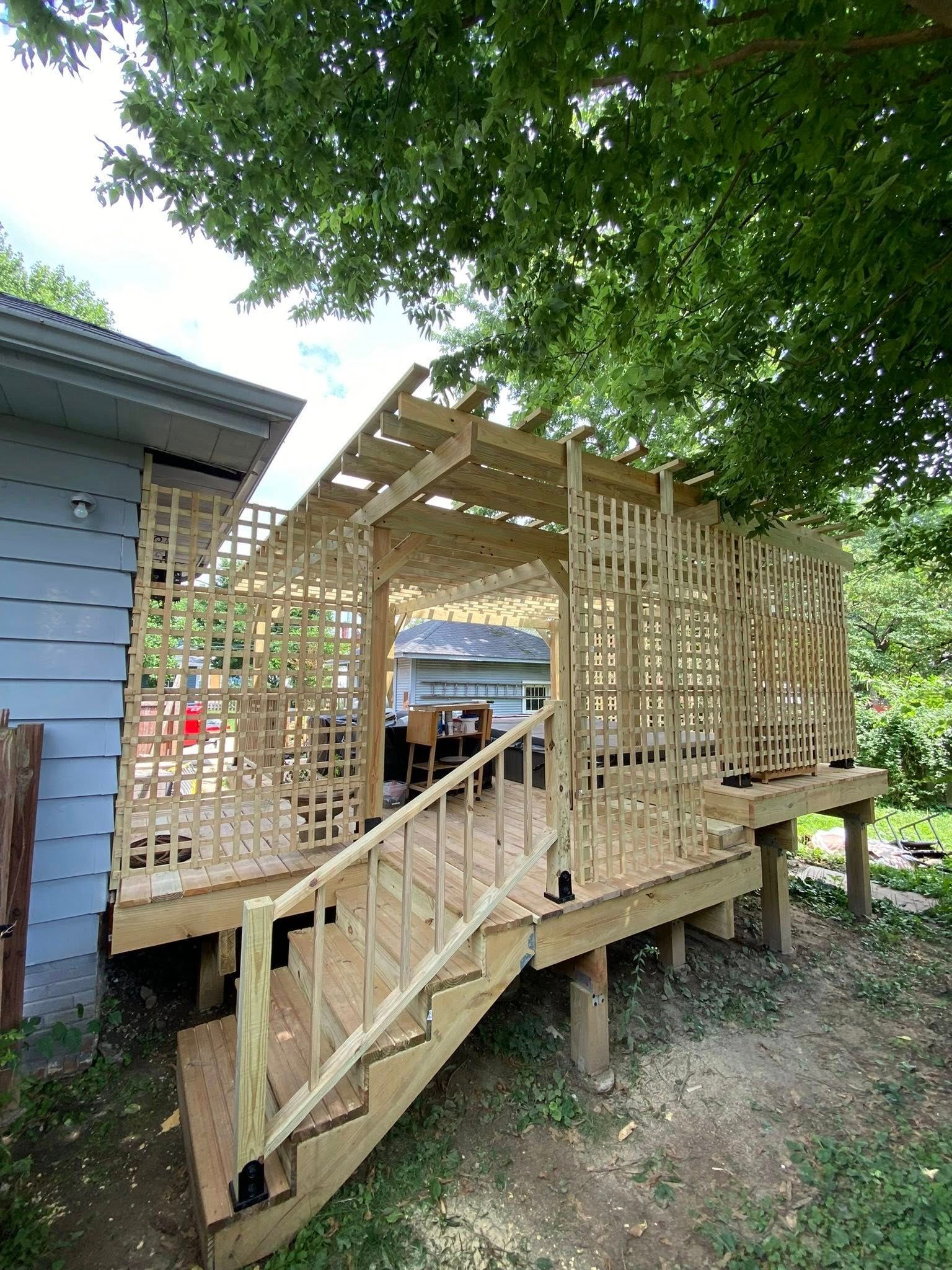 Wooden deck with stairs, trellis, and pergola structure attached to a blue-sided house, under trees.