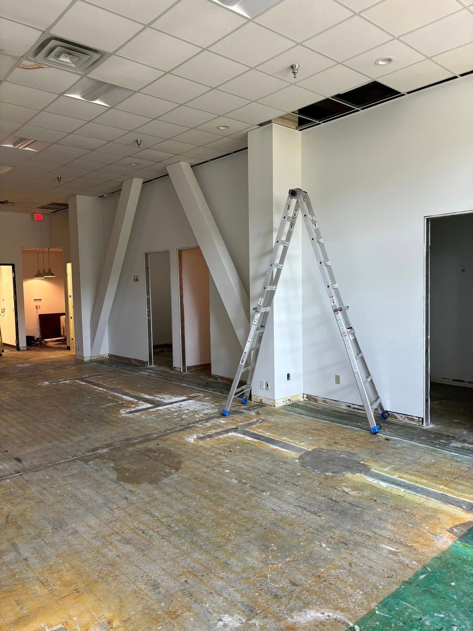 Interior construction site: empty space, ladder propped against wall, exposed flooring, white walls, visible doorways.