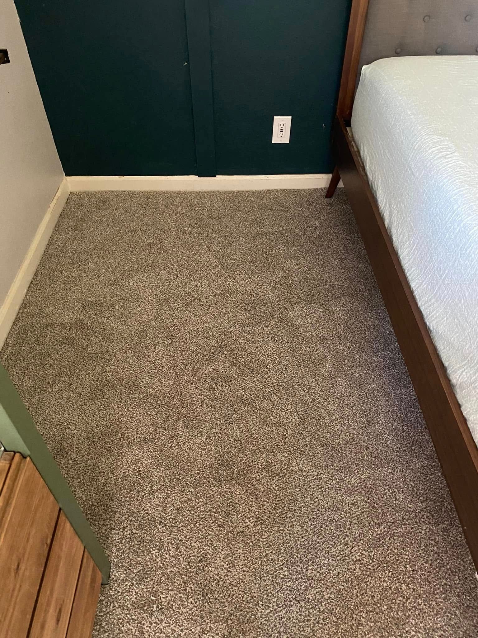 Tan, speckled carpet covers a room floor, alongside a dark green wall and a bed's wooden frame.