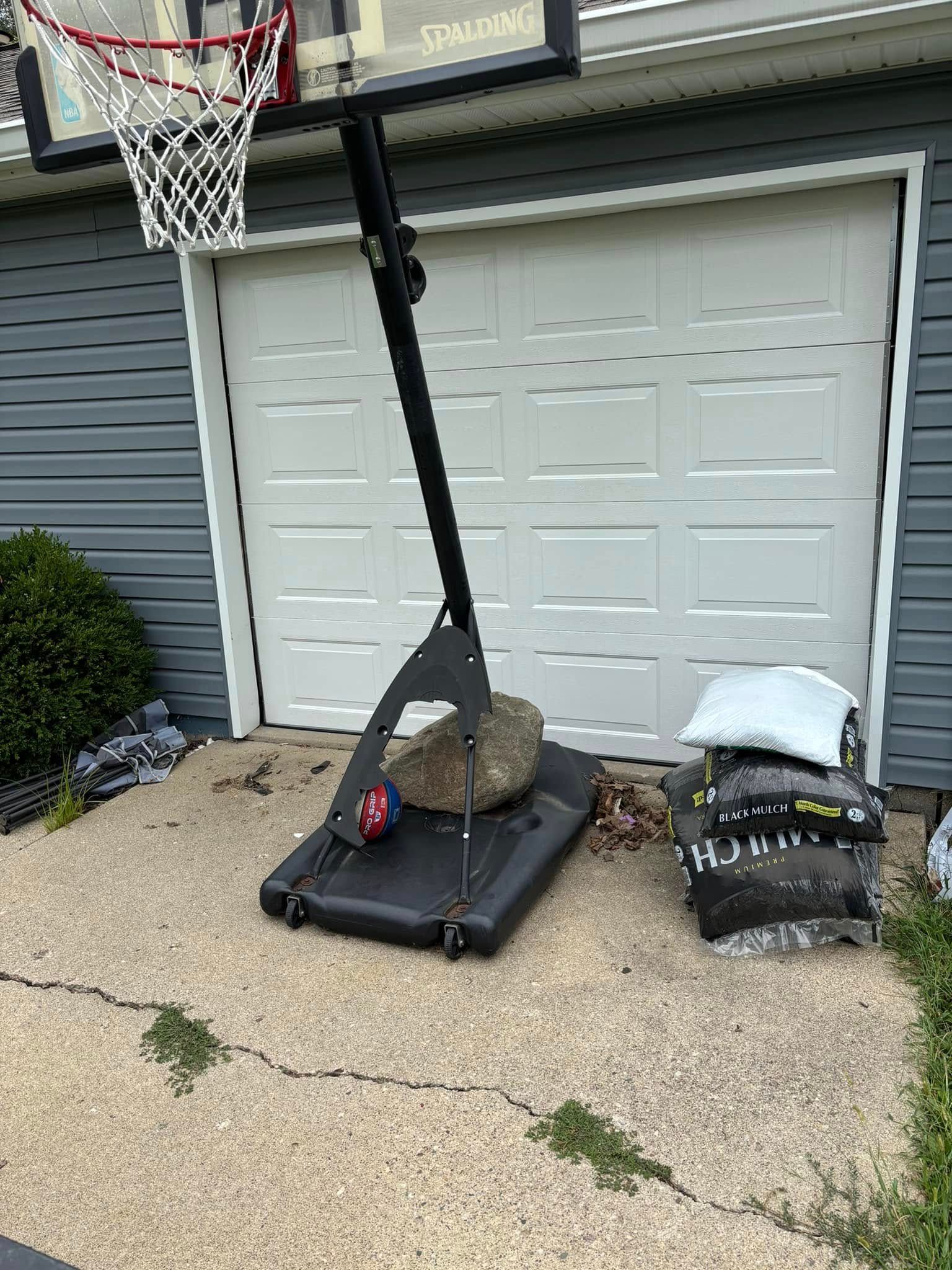 Basketball hoop with a large rock and weights on the base, beside a garage.