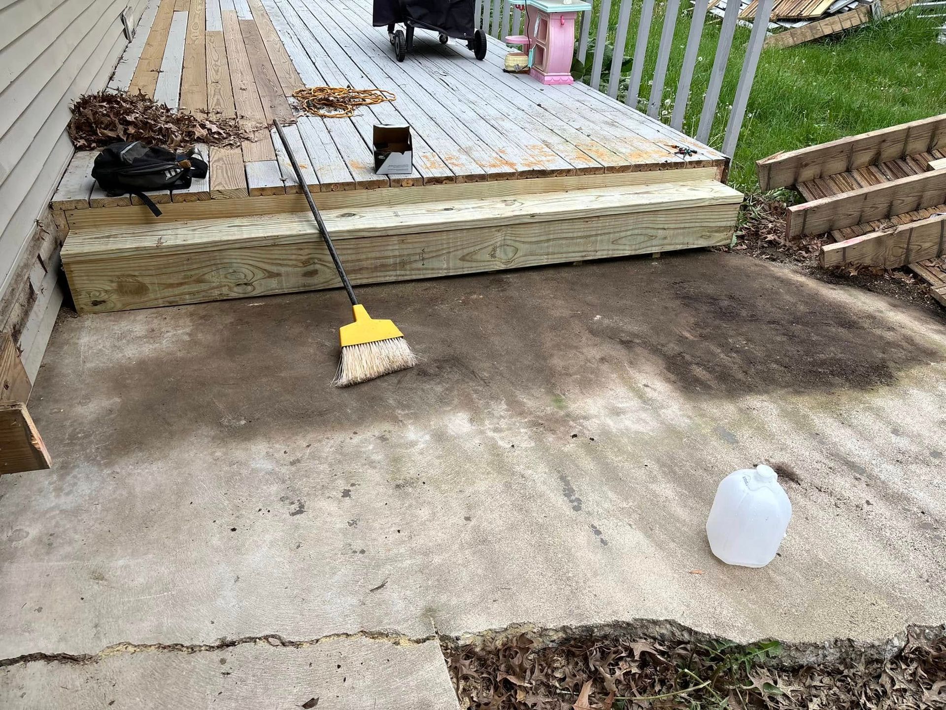 A wooden deck and concrete patio. A broom rests on the patio.