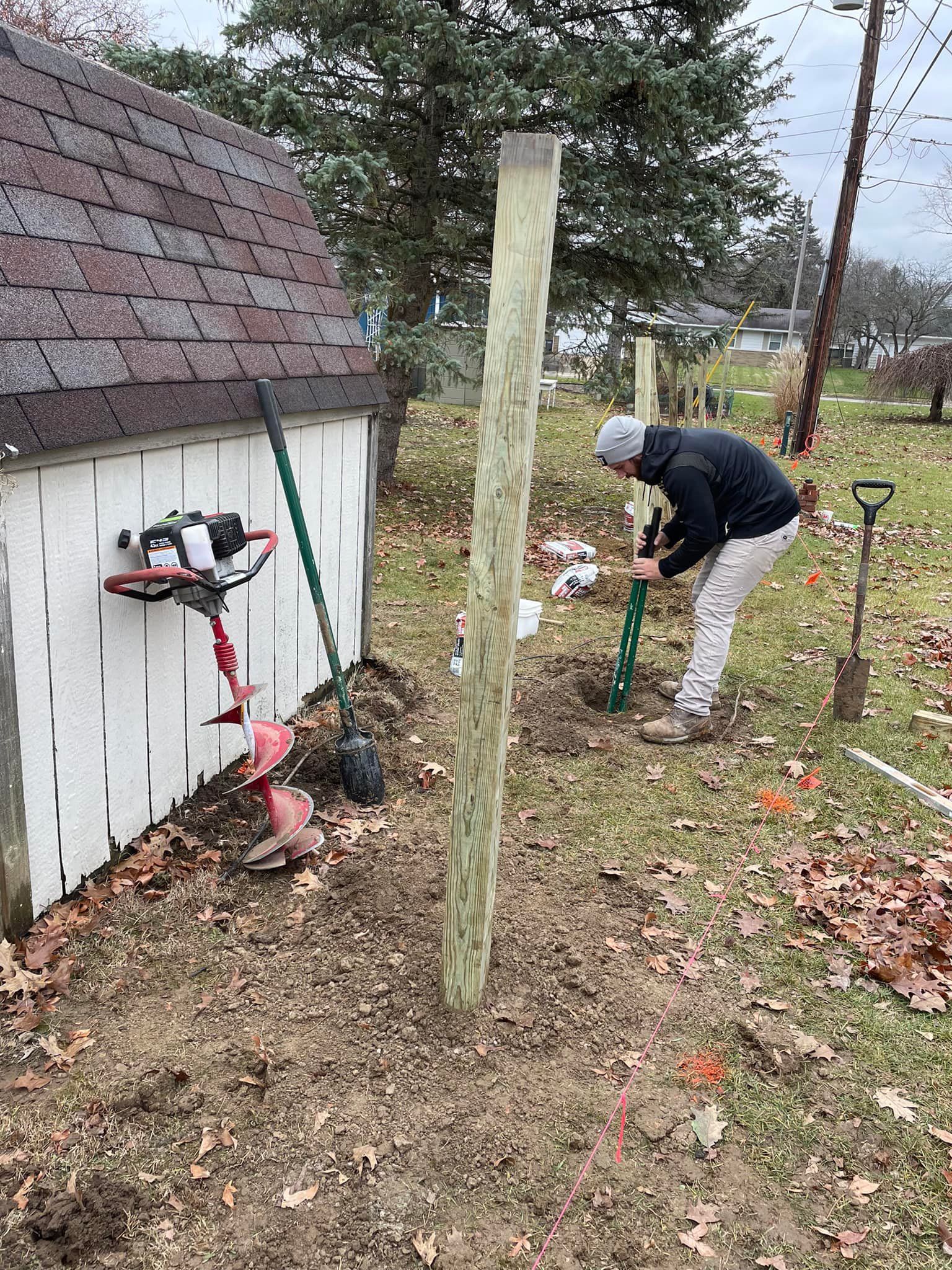 Man installs a fence post. Next to a white shed, using tools on a cloudy day.