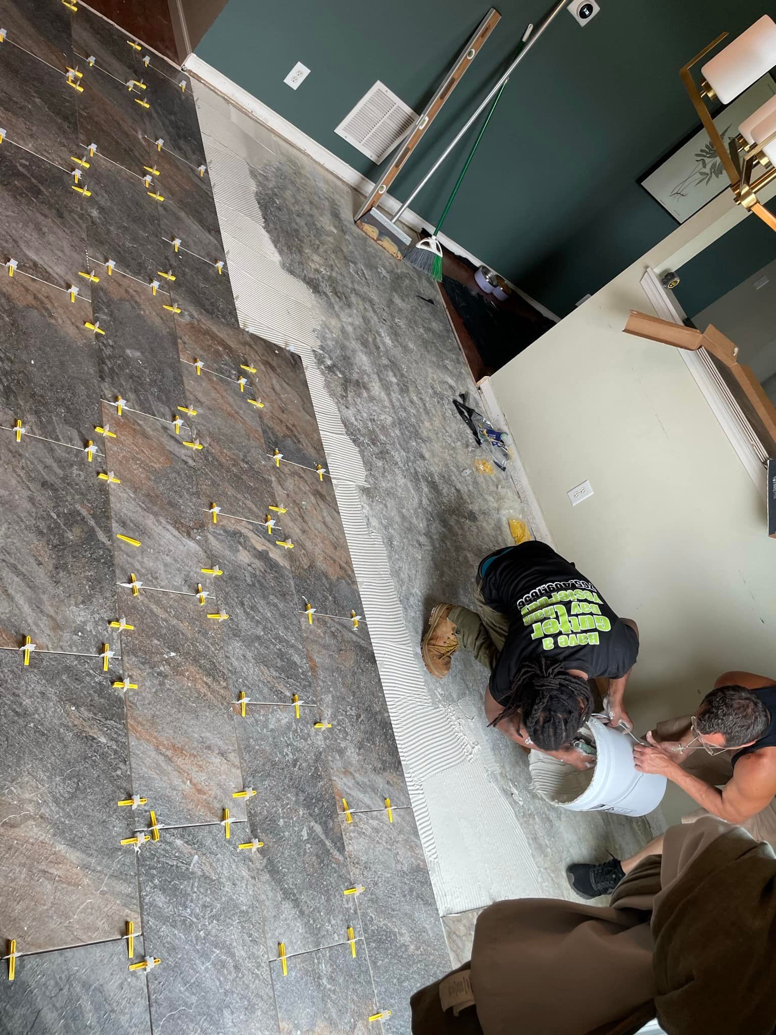 Two people installing tile on a floor; dark brown/gray tile with yellow spacers.